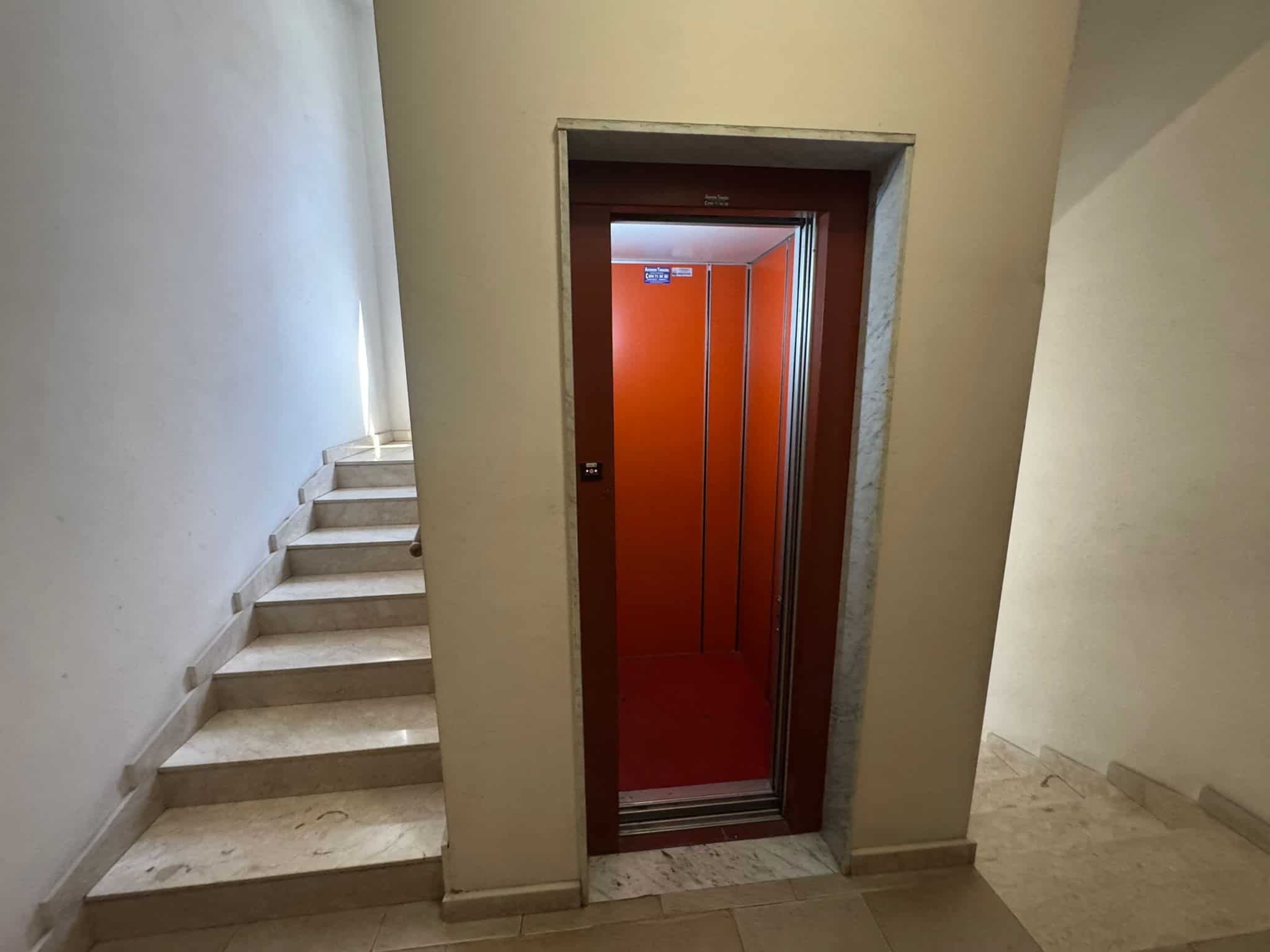 Ascensore in un edificio residenziale con pareti arancioni, situato tra le scale in marmo bianco, pronto per il trasporto di persone tra i piani.