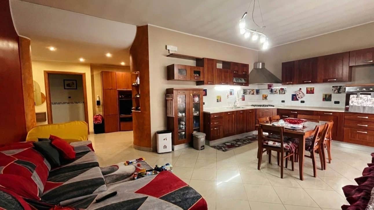 Ampia cucina open space con mobili in legno scuro, zona pranzo e soggiorno con divano colorato, illuminazione moderna, ideale per famiglie e intrattenimento.