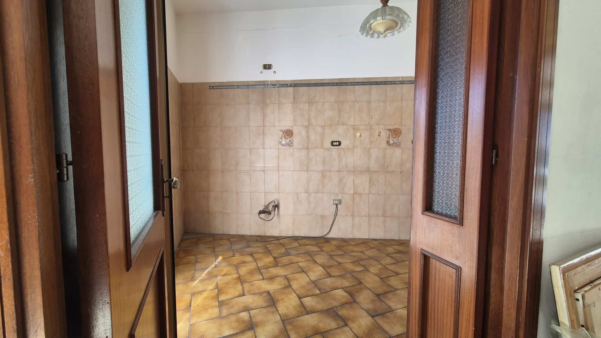 Spazio vuoto in cucina con pareti rivestite di piastrelle beige e pavimento in piastrelle a scacchi marroni e gialle, porte in legno con vetri smerigliati.