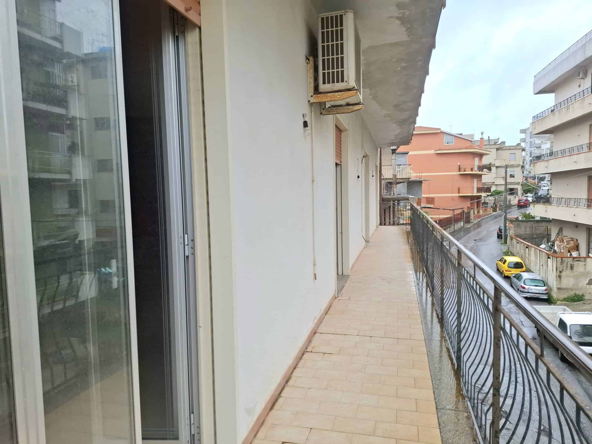 Balcone di un appartamento con vista sulla strada, con ringhiera in ferro battuto e pavimentazione in piastrelle beige, in un quartiere urbano residenziale.