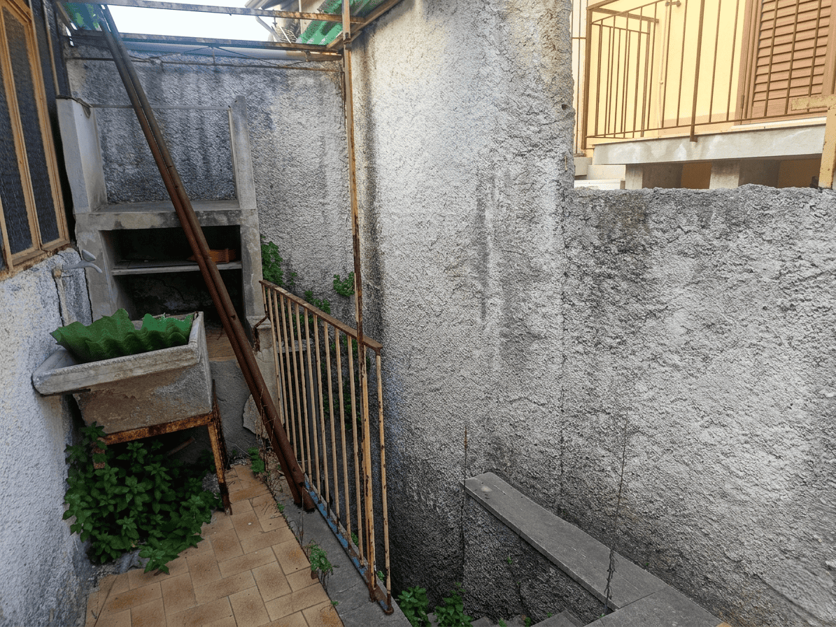 Spazio esterno di un cortile con muri in cemento, ringhiera in metallo e piante verdi, ideale per ristrutturazione o miglioramento abitativo.