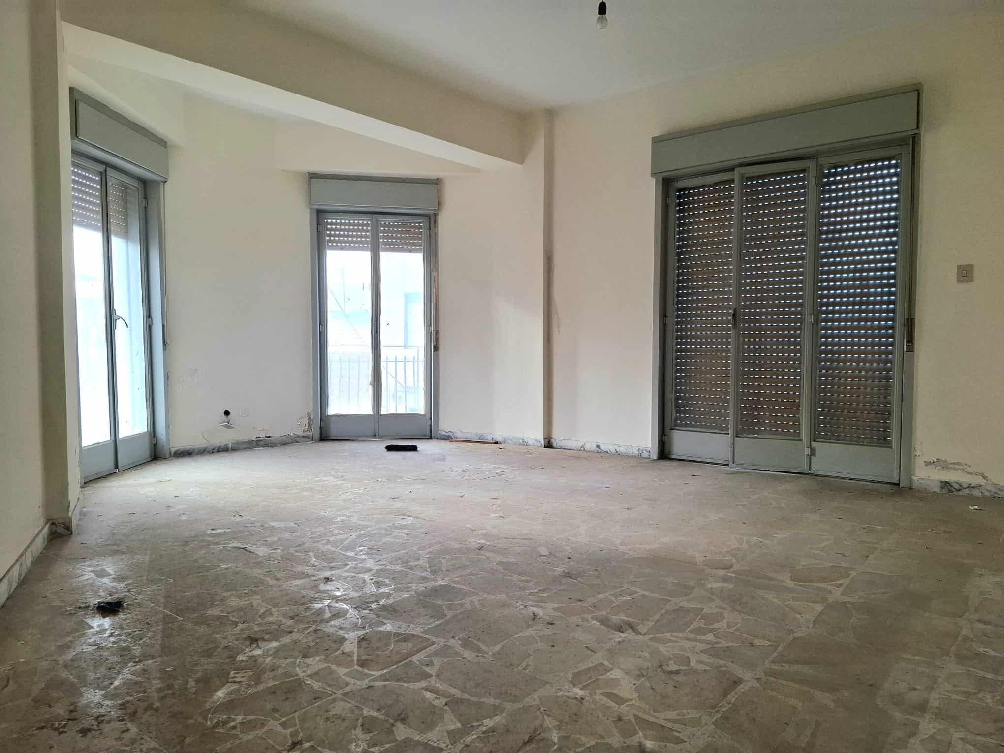 Ampio soggiorno vuoto con grandi finestre e balcone, pronto per essere arredato, in un appartamento in fase di ristrutturazione a NextCasa, ideale per creare uno spazio di comfort e stile.
