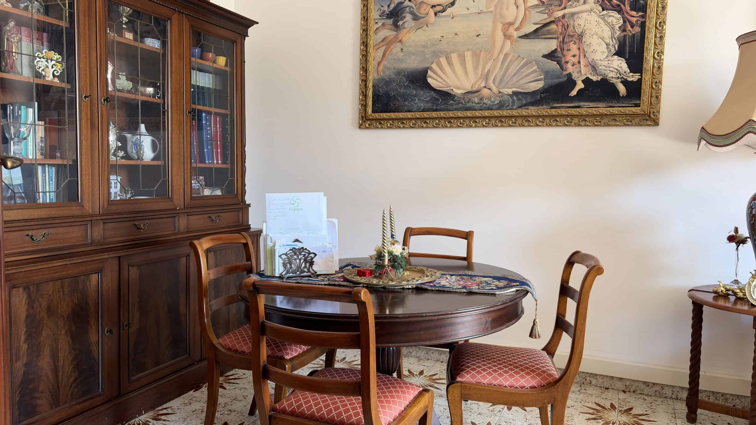 Elegante sala da pranzo con tavolo rotondo in legno e quattro sedie imbottite, arredamento classico con mobile in legno con vetrine e quadro artistico alle pareti, atmosfera accogliente e raffinata.