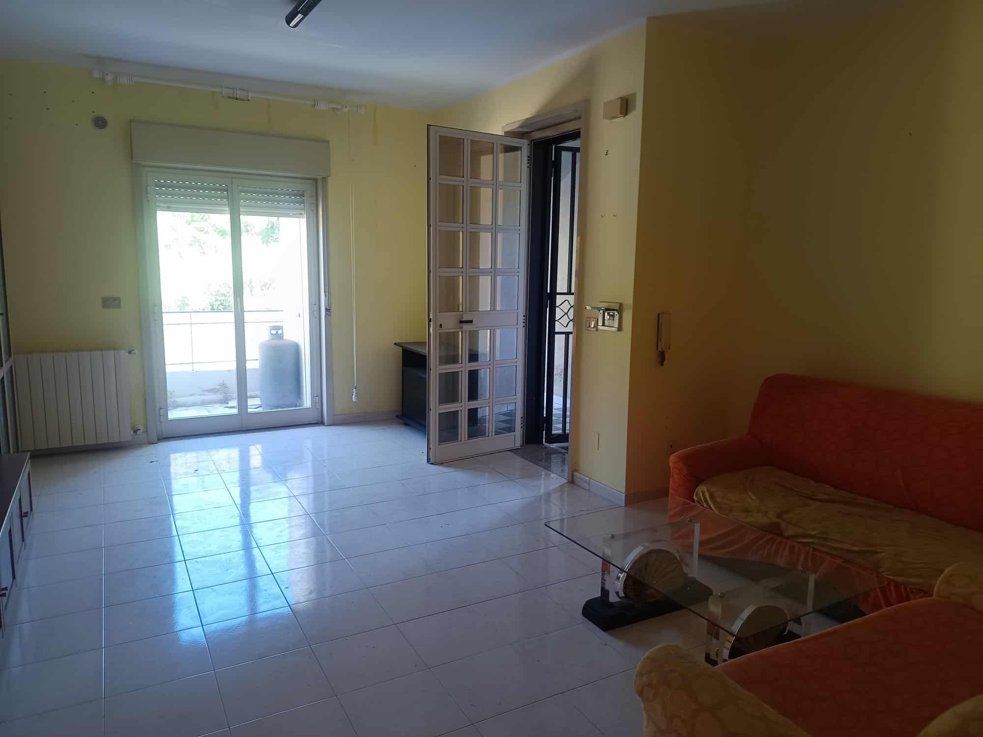 Interno di un appartamento con pareti gialle, porta finestra che dà su un balcone, divano arancione e tavolino in vetro, ambiente luminoso e spazioso.