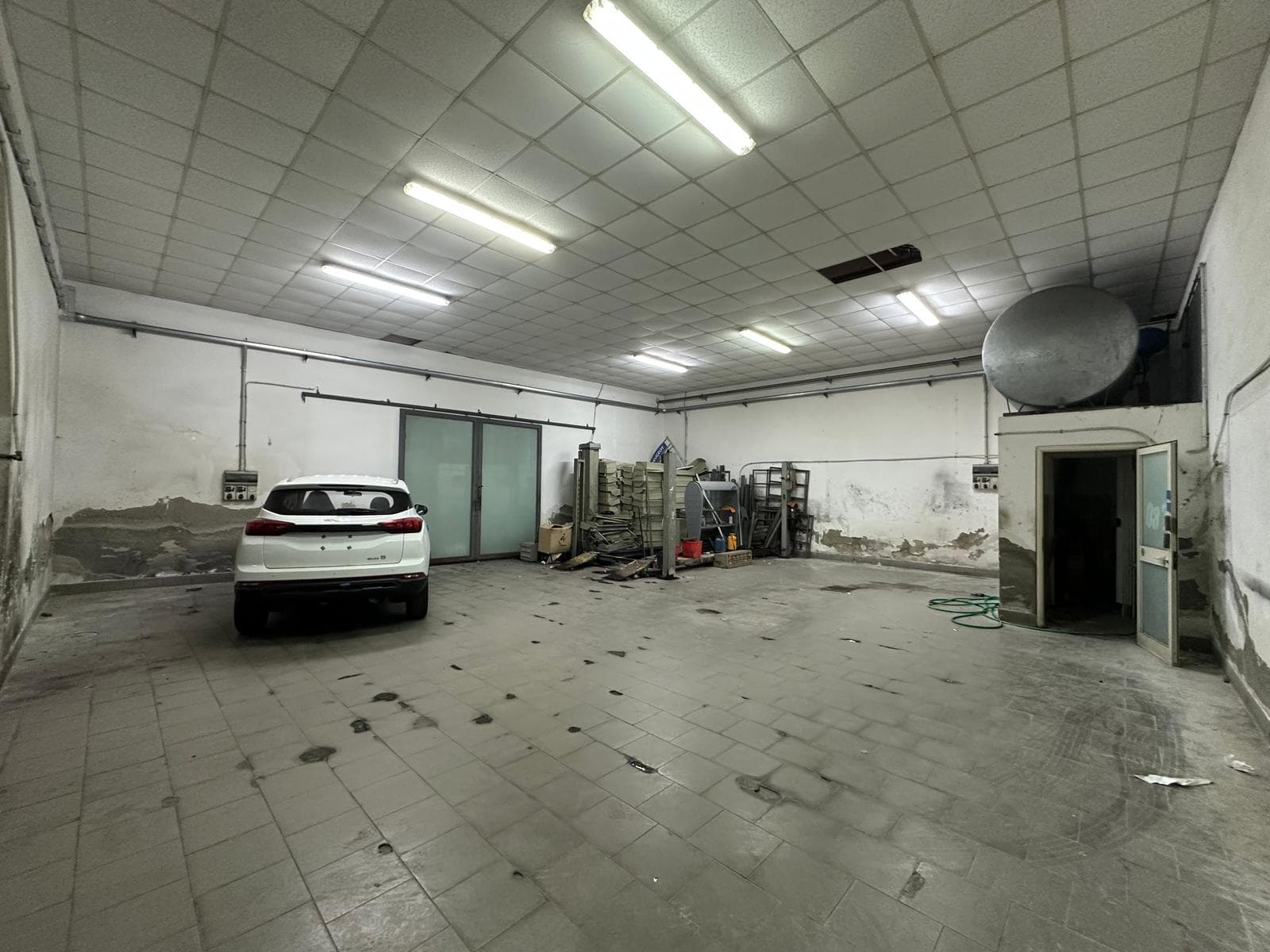 Automobile parcheggiata in un garage industriale vuoto con pareti bianche e pavimento in piastrelle, illuminato da luci fluorescenti.