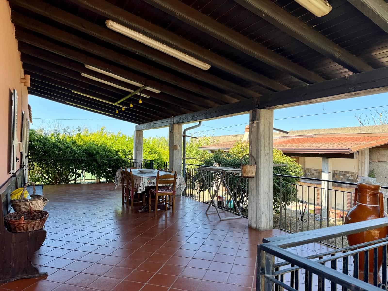 Ampia terrazza coperta con arredi in legno, vista su giardino e case vicine, ideale per relax e cene all'aperto, con pavimentazione in piastrelle rosse e ambiente luminoso.