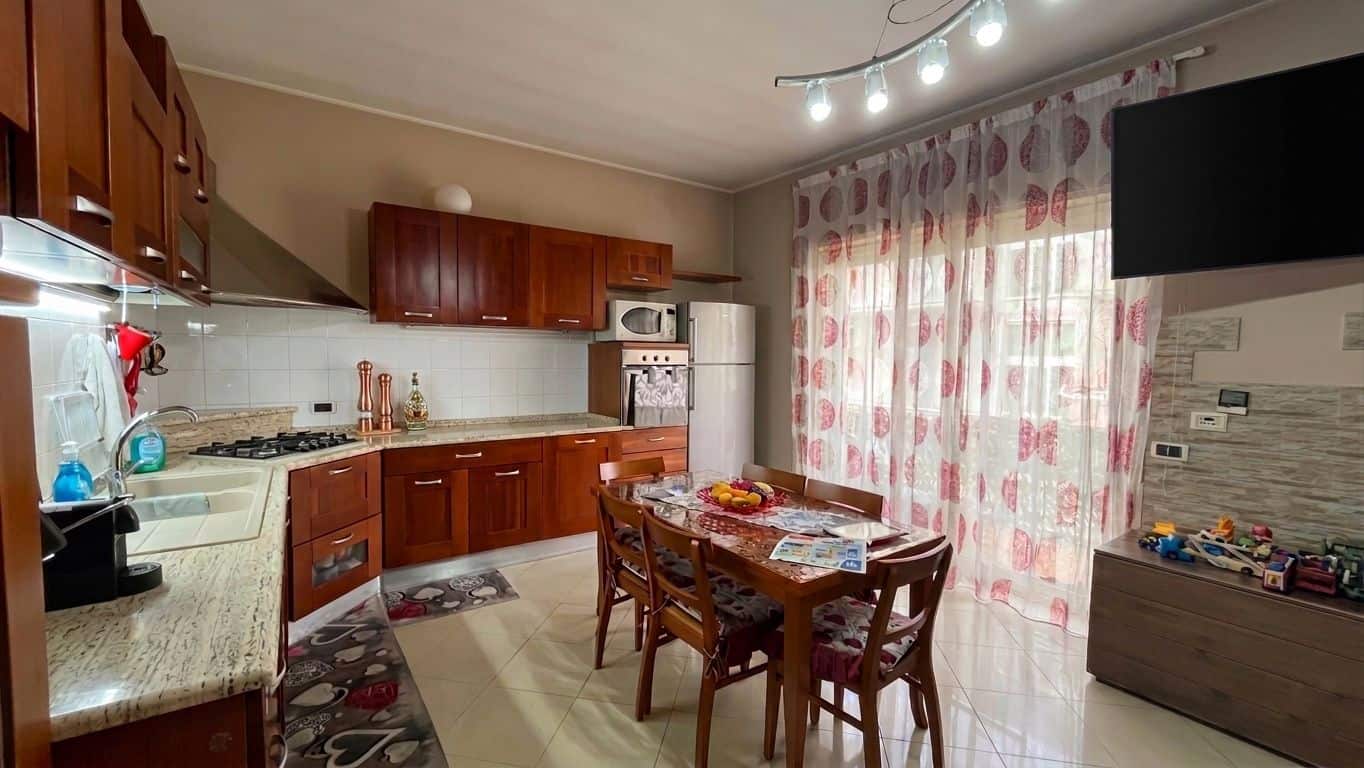Piano cucina moderno con mobili in legno scuro, tavolo da pranzo, elettrodomestici e grande finestra con tende a motivi floreali, ambiente luminoso e accogliente.