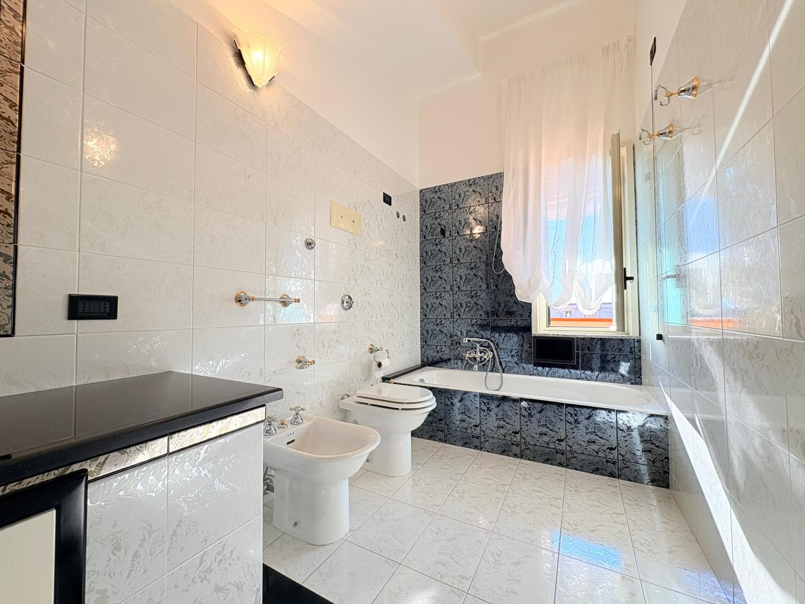 Lavabo e bidet in un bagno spazioso con pareti e pavimenti in piastrelle bianche e nere, finestra con tende leggere, illuminazione soffusa, ideale per ristrutturazioni e arredi moderni.