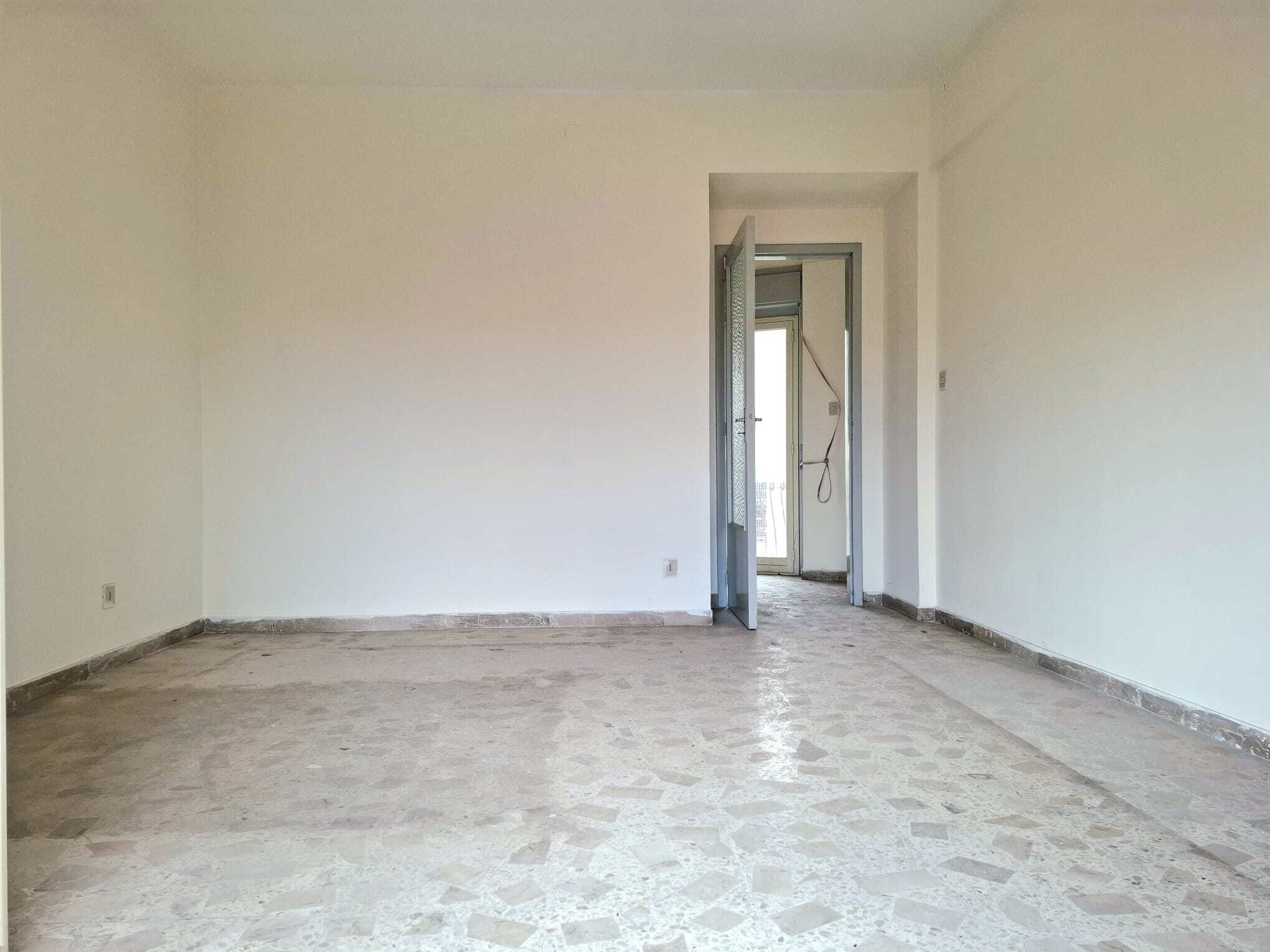 Ampia stanza vuota con pareti bianche e pavimento in piastrelle, con porta finestra che conduce a un balcone, pronta per essere arredata e personalizzata.