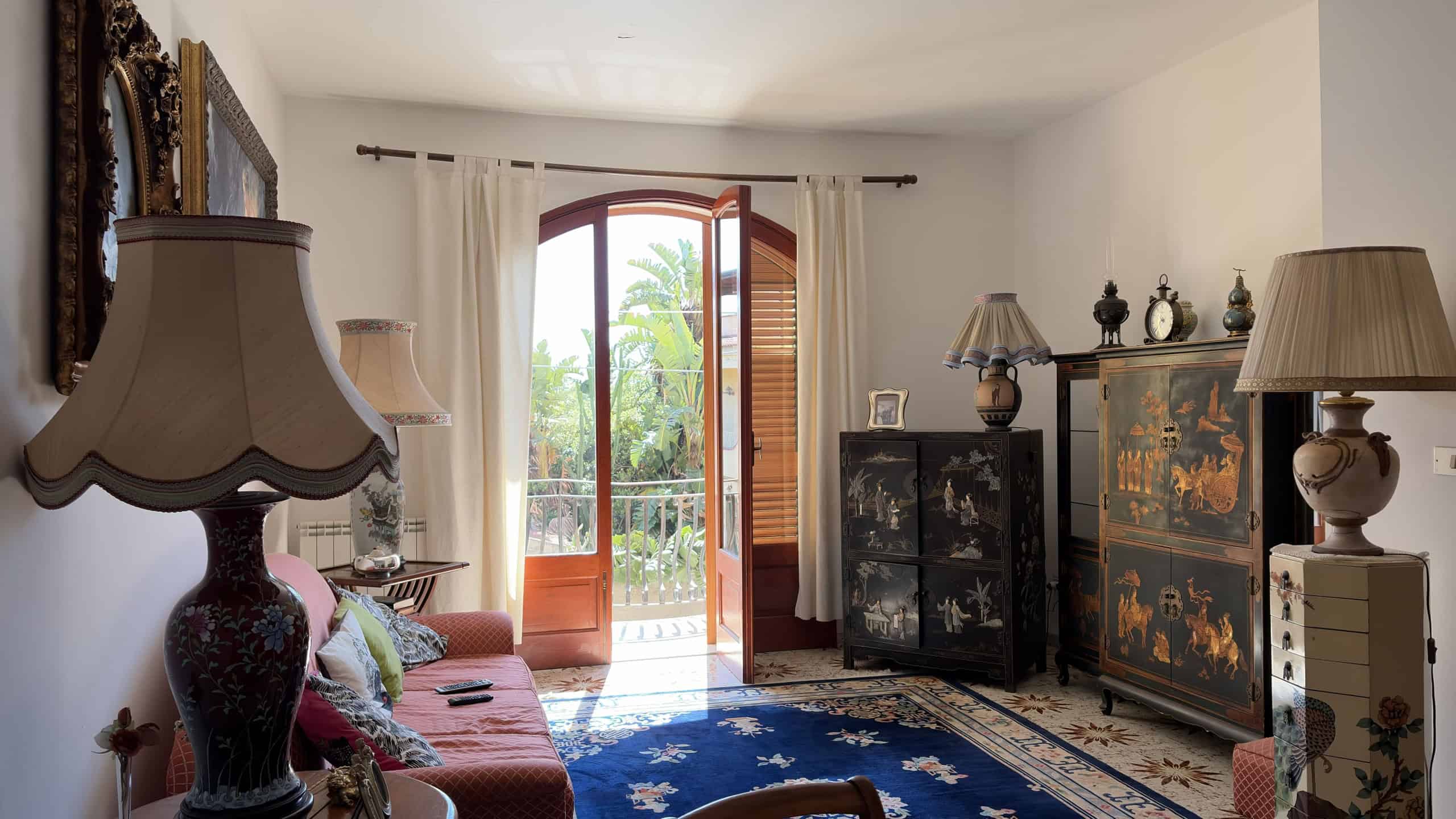 Elegante soggiorno con porta finestra che si apre su un balcone, arredato con mobili classici, lampade e tappeto blu, ideale per ambienti accoglienti e raffinati.