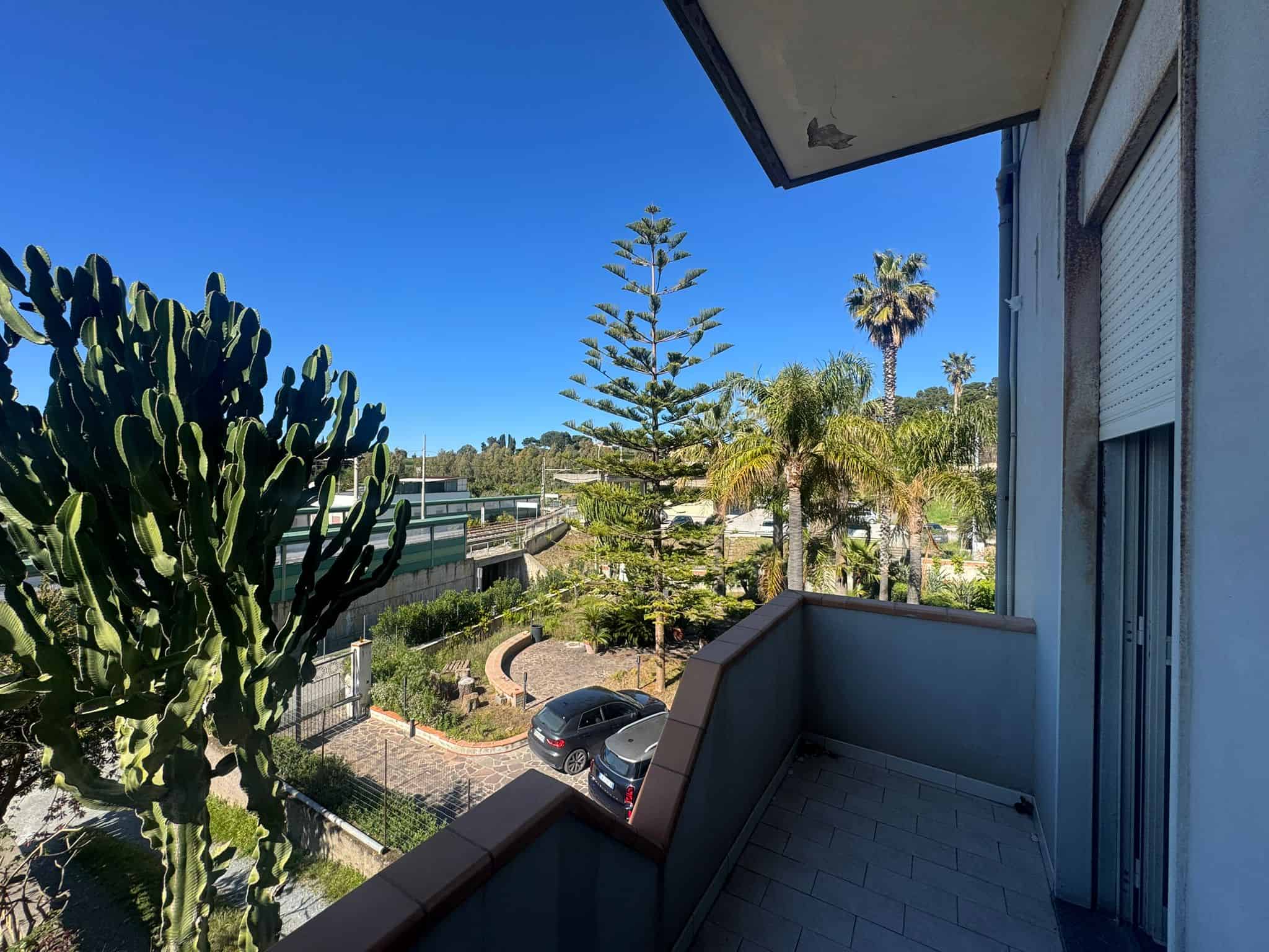 Ampio balcone con vista su giardino e strada, circondato da piante tropicali e alberi alti, ideale per relax e momenti all'aperto in un contesto residenziale tranquillo.