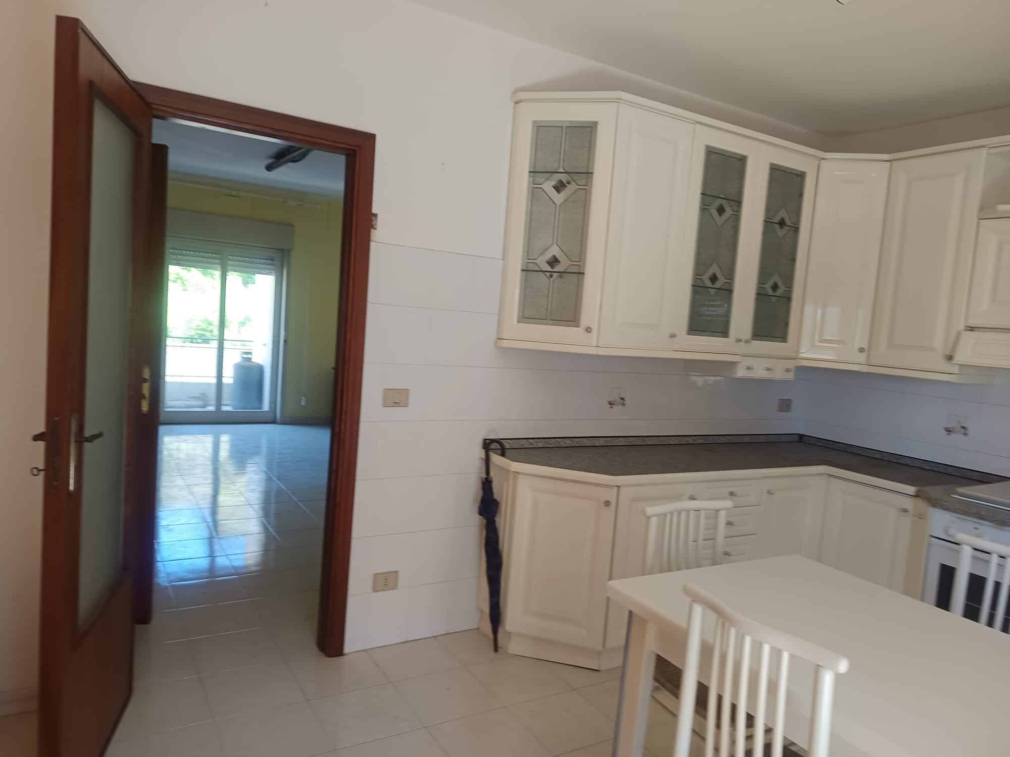 Ampia cucina moderna con mobili bianchi, piano in granito e accesso a un soggiorno luminoso con grandi finestre e balcone, ideale per vivere e cucinare in comfort.