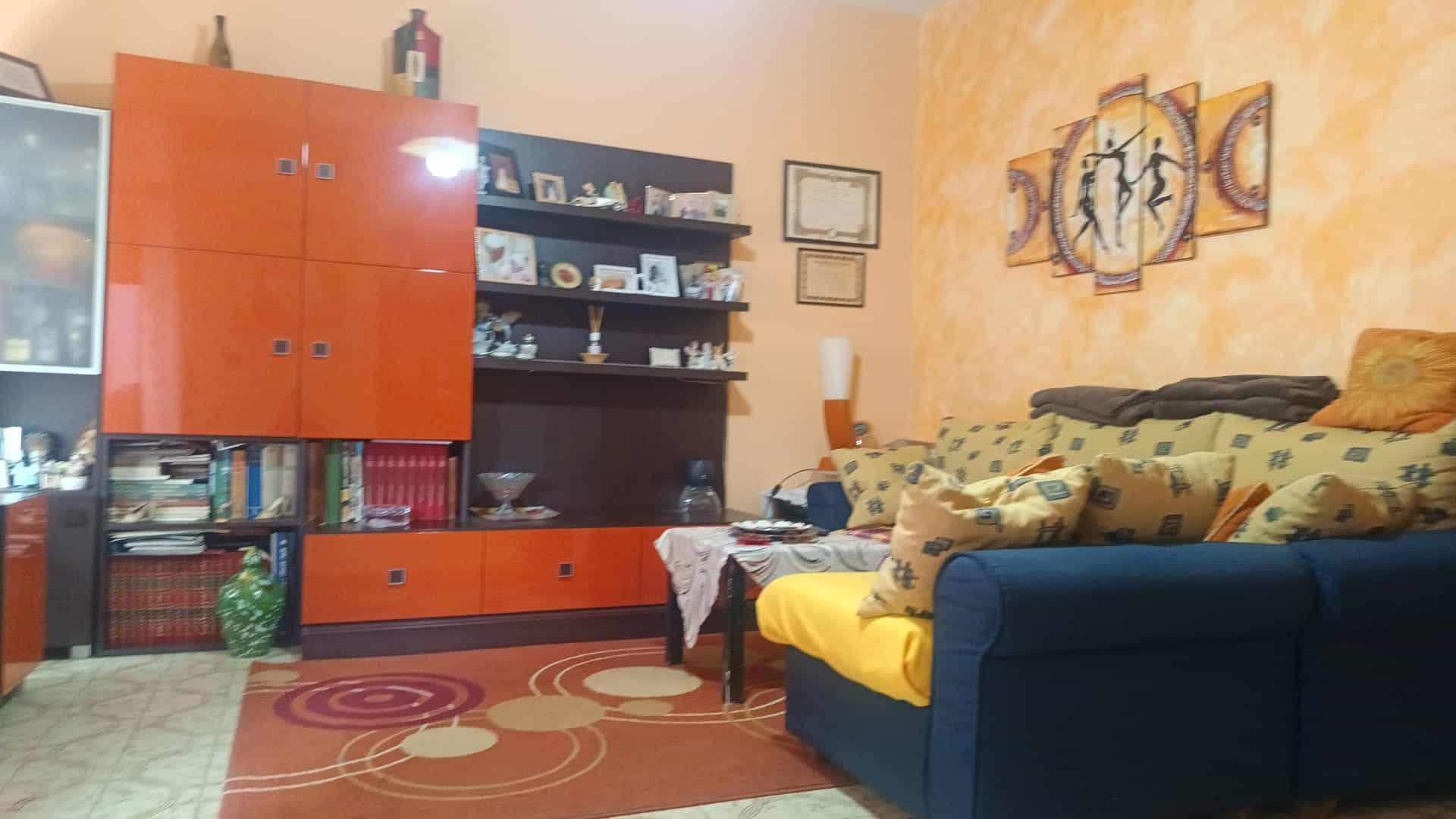 Ampia zona living con divano colorato, libreria con oggetti decorativi e pareti arancioni, ideale per arredamento di case moderne e accoglienti.