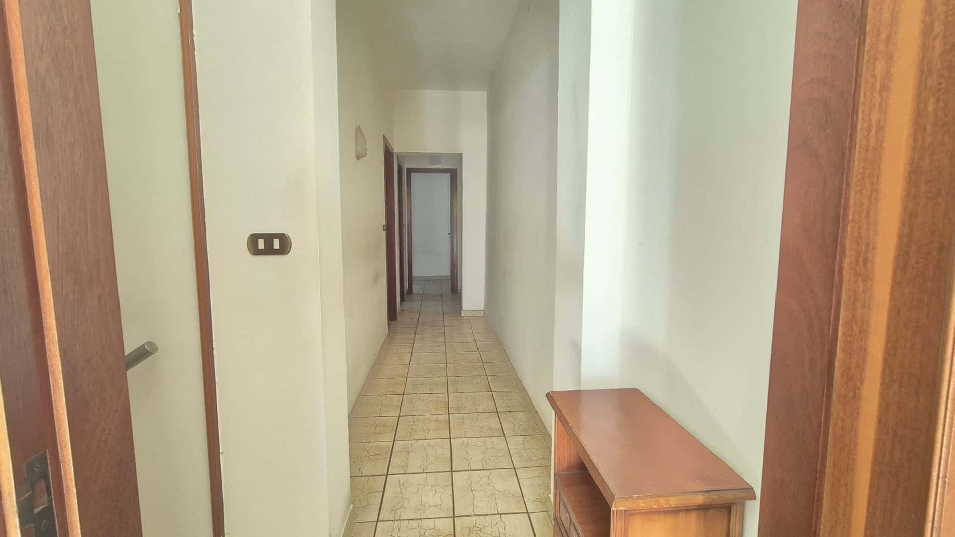 Ampia corridoio interno di un appartamento con pareti bianche e pavimento in piastrelle beige, vista verso le porte delle stanze e l'ingresso, ambiente luminoso e ben tenuto.