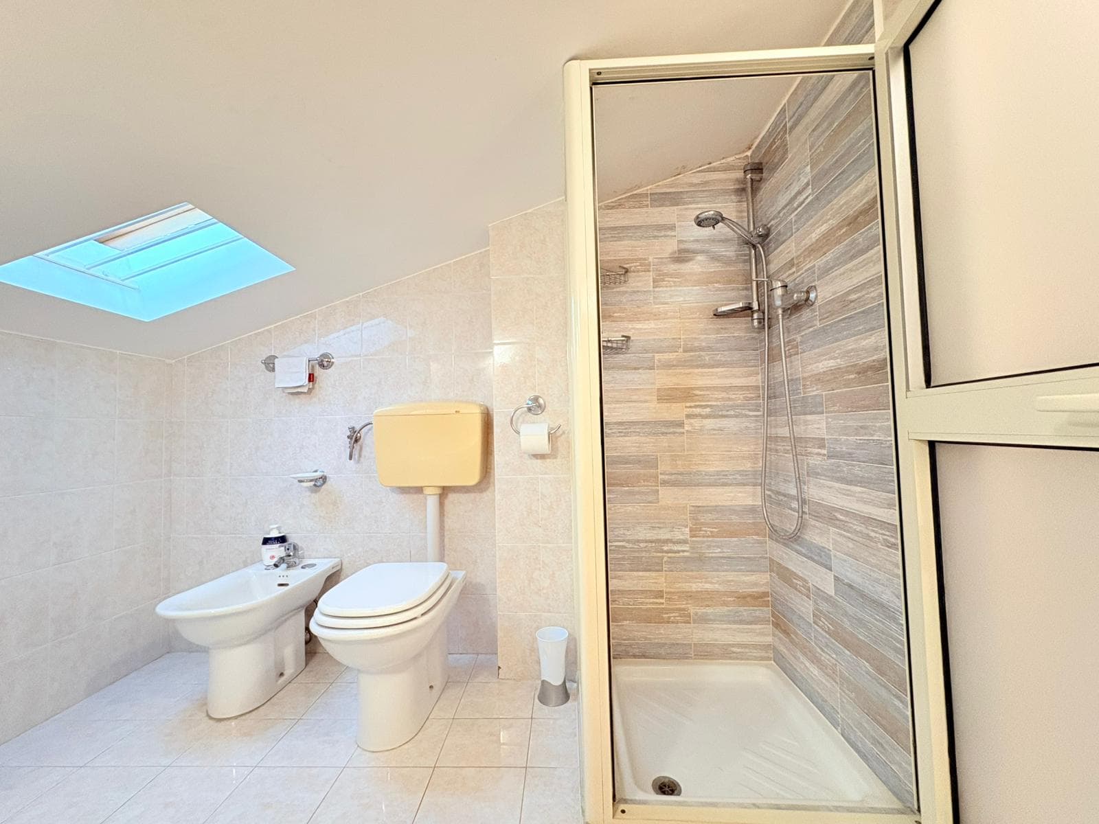 Doccia moderna con pareti in piastrelle effetto legno, bagno spazioso con WC e bidet, illuminato da luce naturale tramite finestra e lucernario sul soffitto.