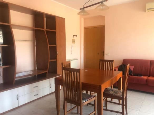 Interno soggiorno con tavolo in legno, divano rosso e mobile con scaffali, ambiente luminoso e accogliente.