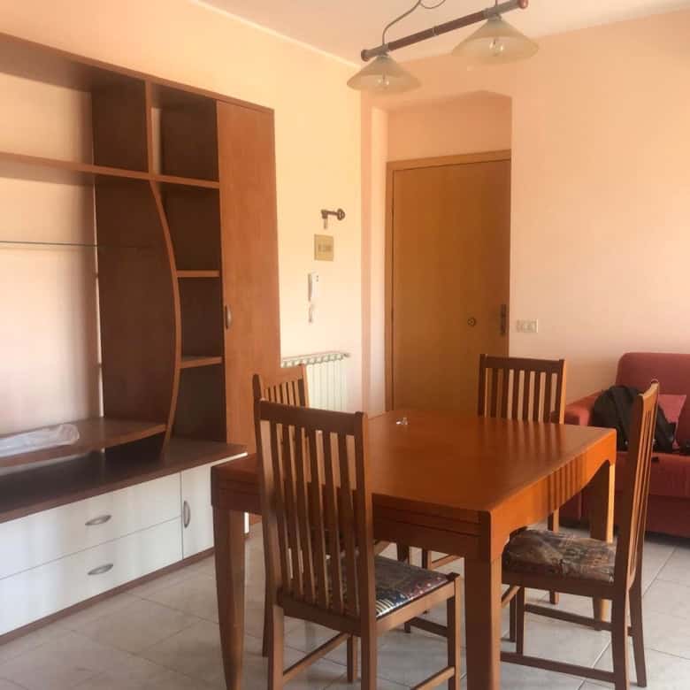 Interno soggiorno con tavolo in legno, divano rosso e mobile con scaffali, ambiente luminoso e accogliente.