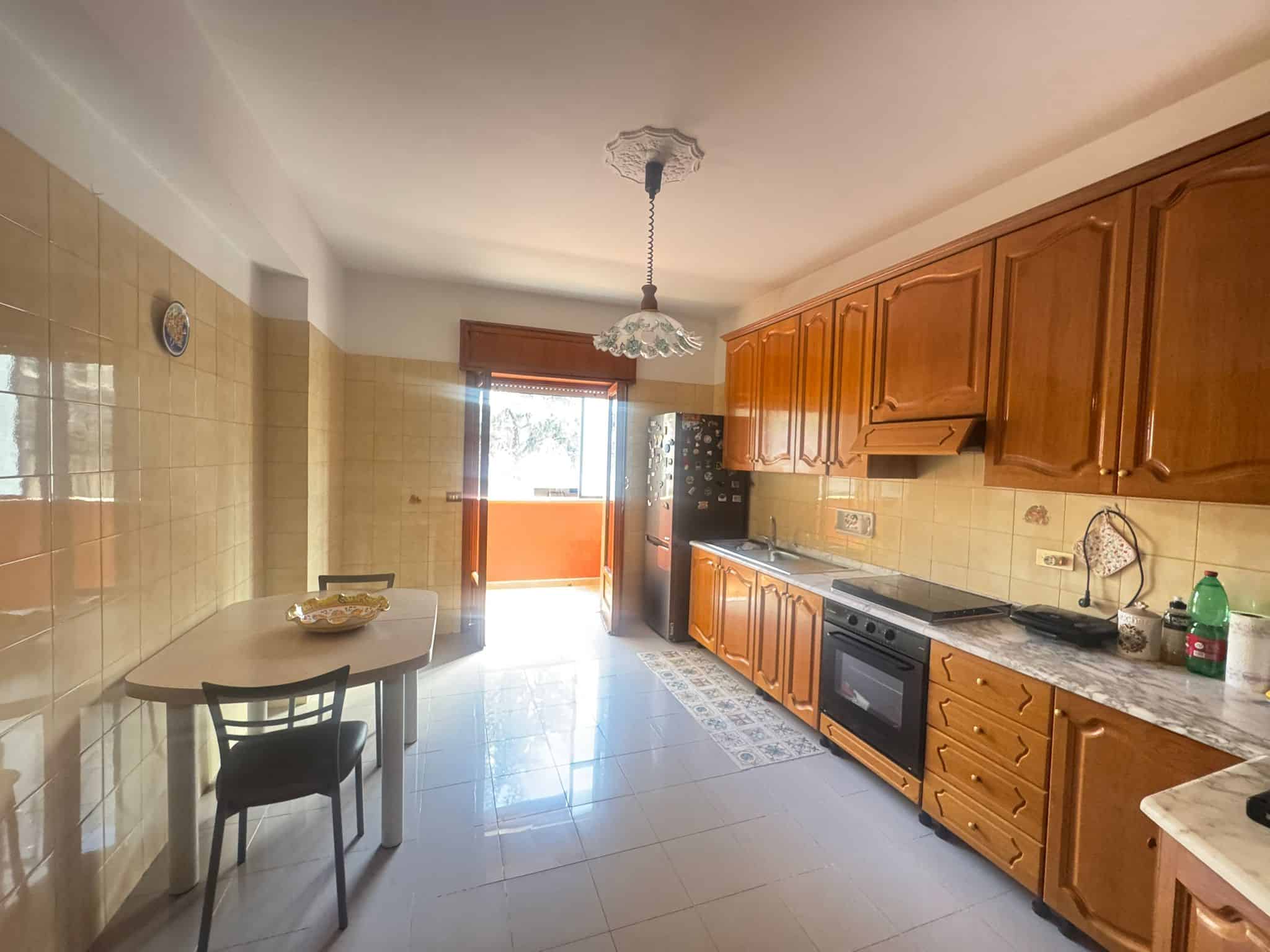 Una cucina spaziosa con mobili in legno, piano cottura e frigorifero, illuminata dalla luce naturale proveniente da una porta finestra con tende in tessuto. Ideale per vivere in comfort e stile.