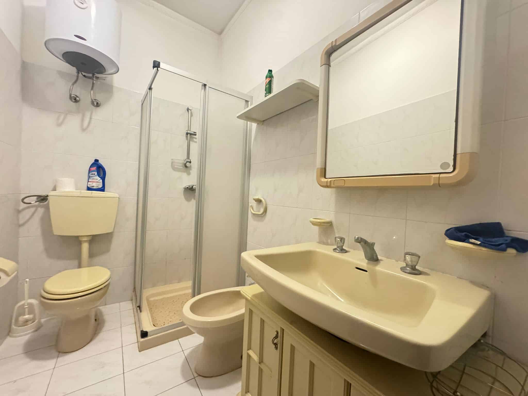 Lavabo e servizi igienici in un bagno semplice con doccia, specchio e accessori essenziali, in un ambiente funzionale e pratico. Ideale per spazi di piccole dimensioni o ristrutturazioni rapide.