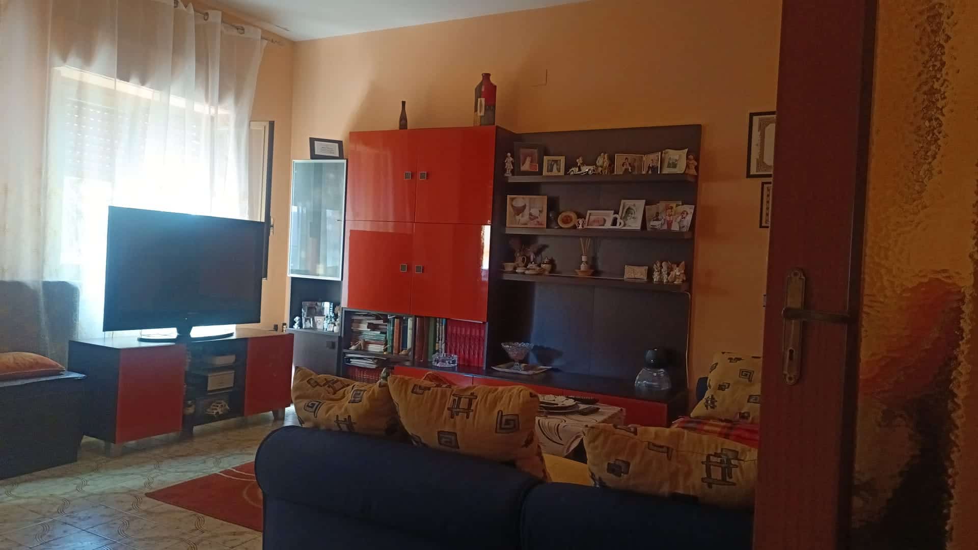 Ampia zona living con divano, televisore e mobili moderni, luminosa grazie a grandi finestre con tende leggere, ideale per vivere e rilassarsi in un ambiente accogliente e funzionale.