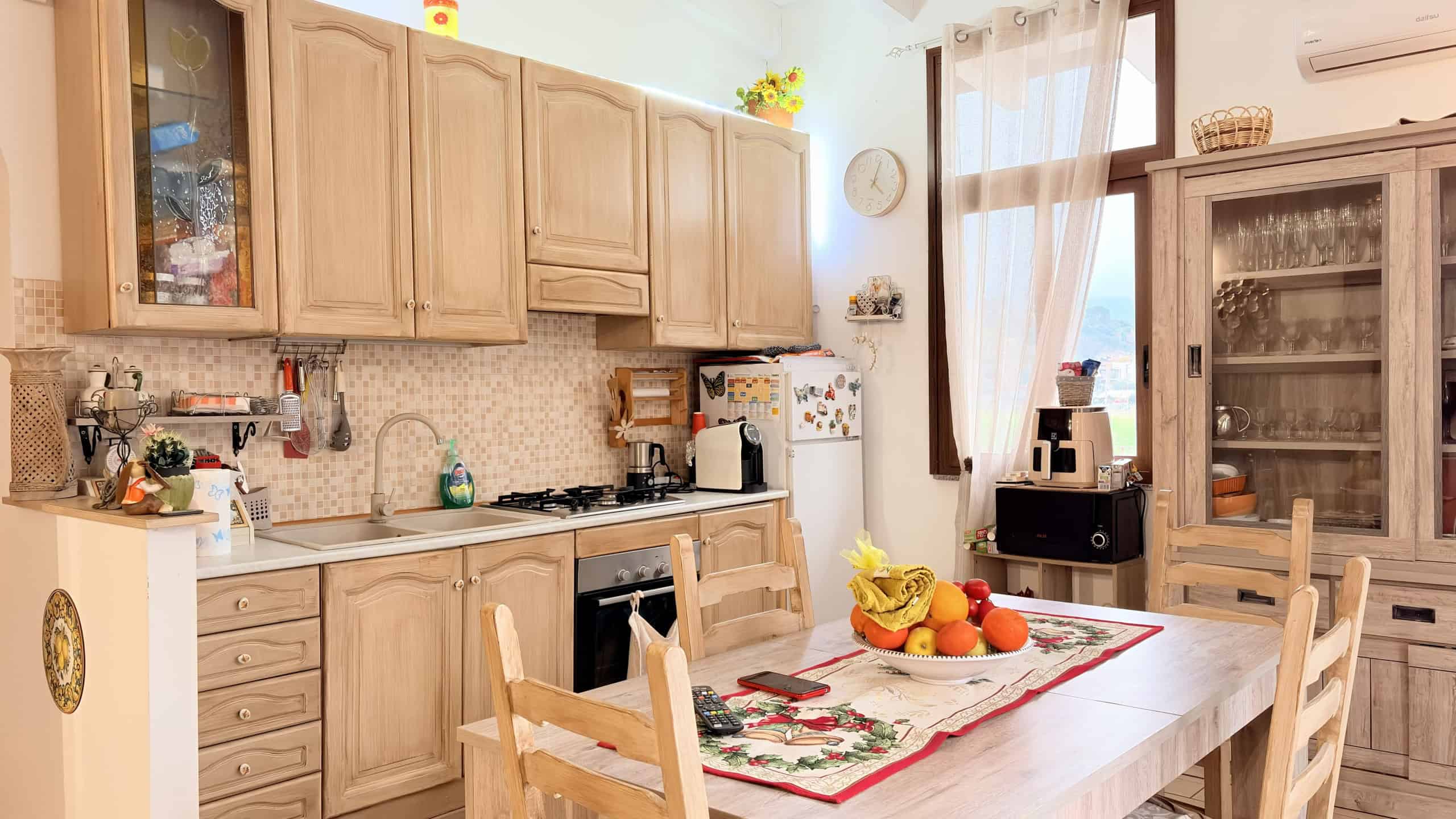 Pannello della cucina in legno chiaro con tavolo da pranzo, frutta fresca e accessori da cucina, ambiente luminoso e accogliente con grandi finestre e tende leggere, ideale per la vita quotidiana e mo.
