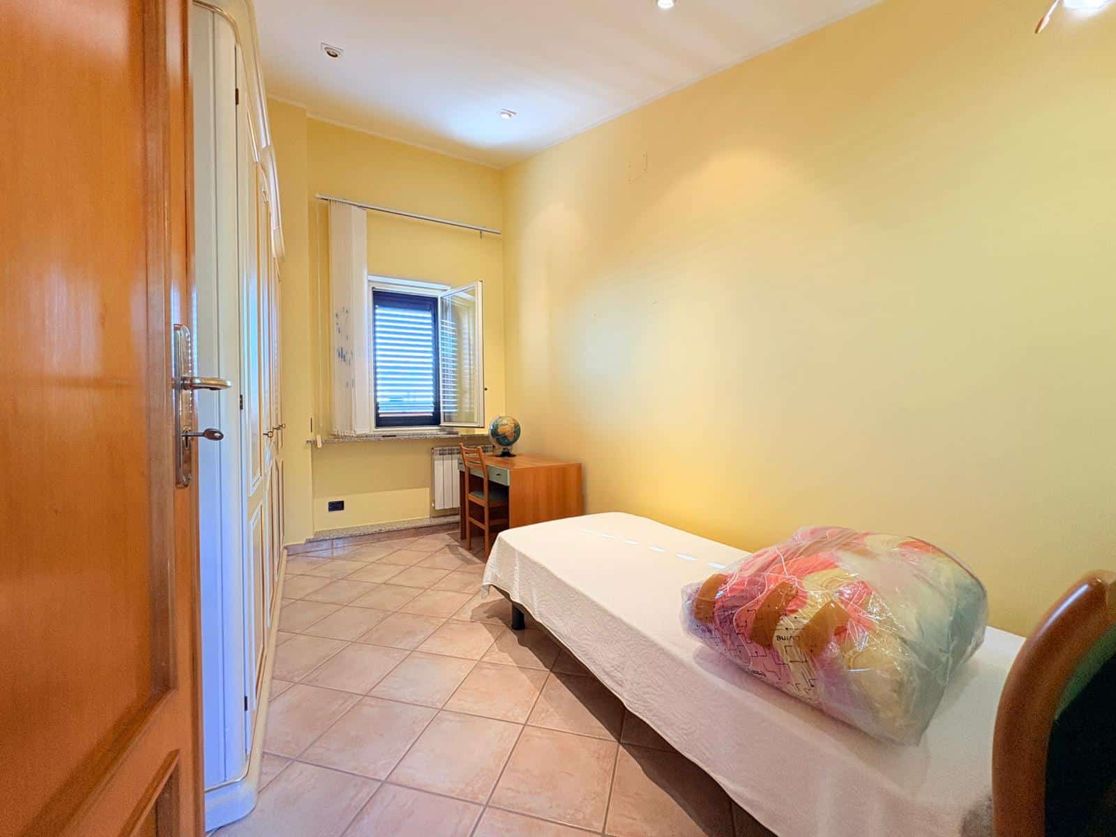 Letto singolo in camera luminosa con pareti gialle, finestra con persiana e arredi semplici, ideale per camere da letto in appartamenti o case private.