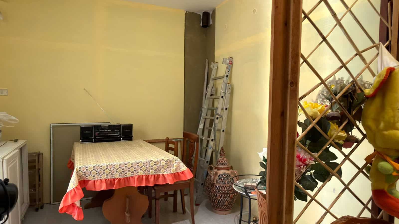 Tavolo da pranzo in soggiorno con sedie in legno, decorazioni e scaffale con oggetti vari, ambiente accogliente e luminoso, ideale per vivere momenti conviviali.