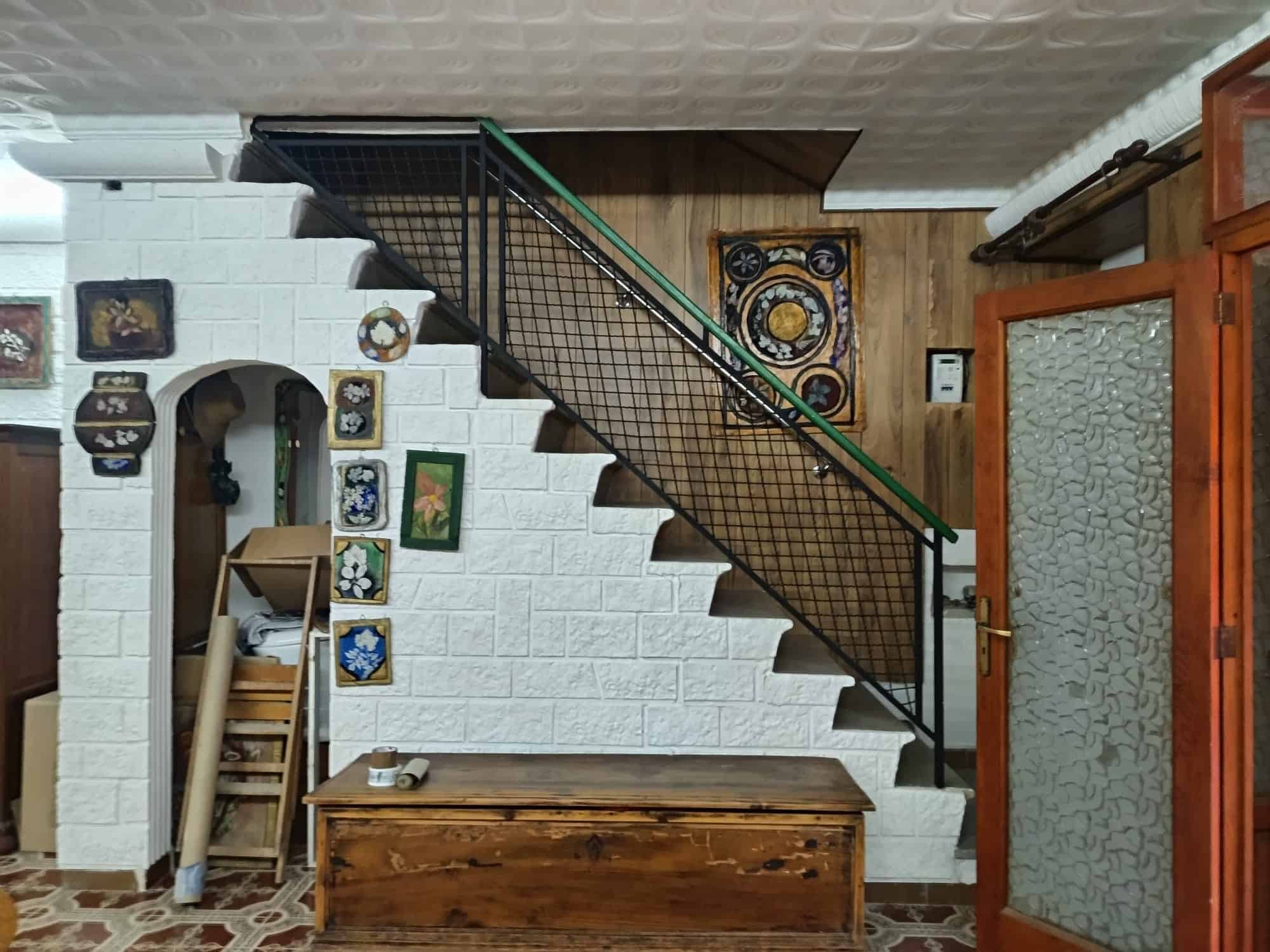 Panca in legno sotto la scala con decorazioni vintage e pareti bianche in mattoni, ambiente accogliente e rustico, ideale per ingressi o zone di passaggio in case di charme.
