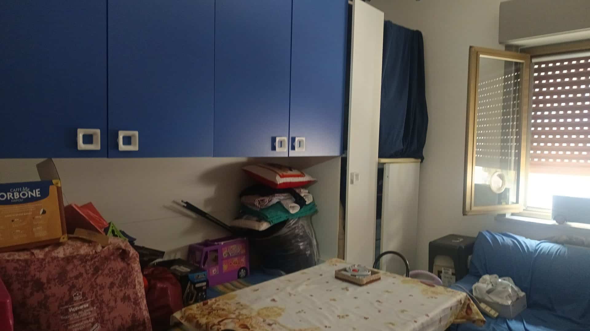 Armadio a muro blu con ante a battente in una stanza domestica, con oggetti e scatole sul tavolo e vicino alla finestra aperta.