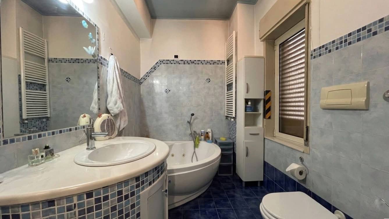Lavabo e vasca da bagno in un bagno moderno con piastrelle azzurre e bianche, finestra con tapparella, accessori bagno e asciugamani.
