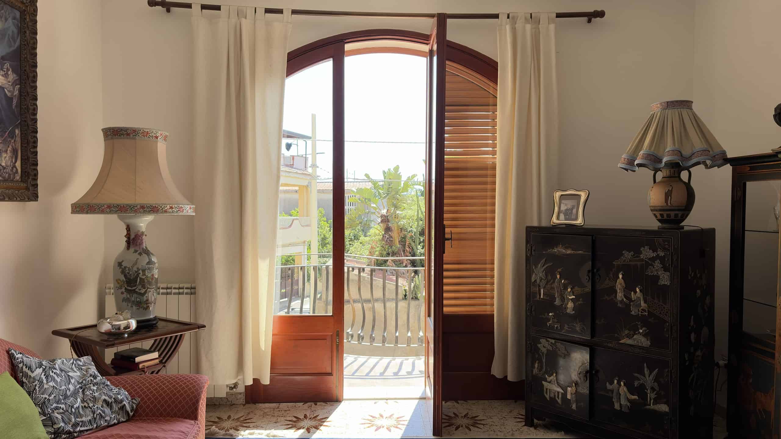 Elegante porta finestra in legno con tende bianche, che si apre su un balcone con vista su alberi e case, in un soggiorno classico arredato con lampade e mobili decorativi.