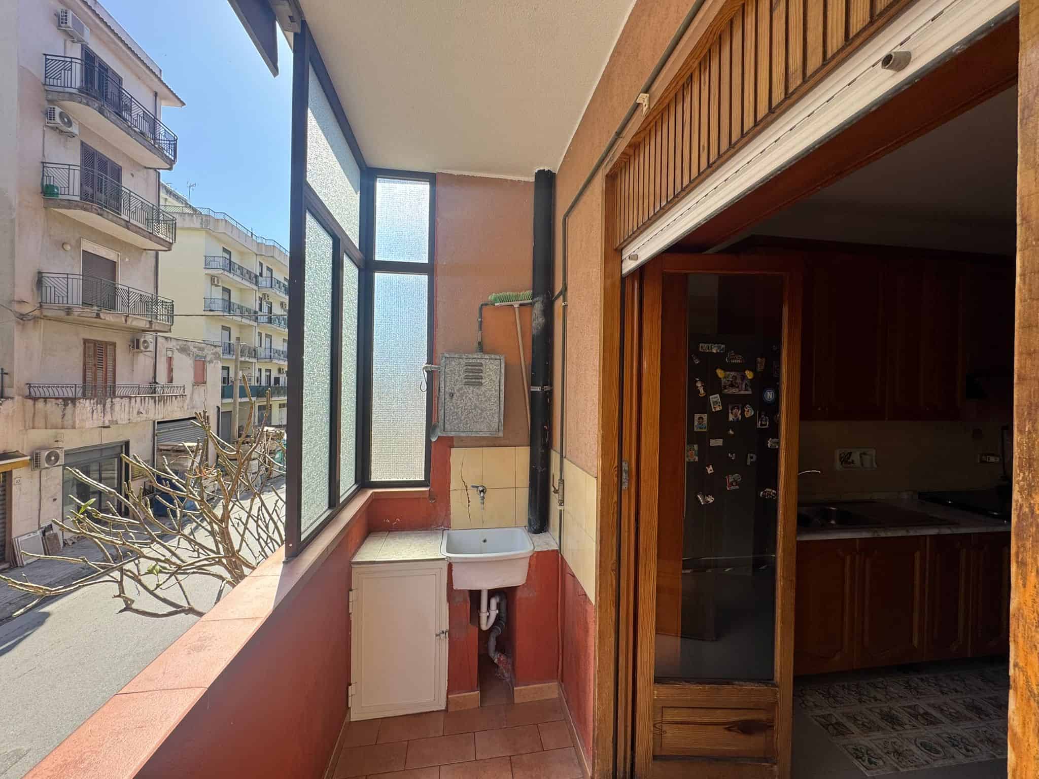 Lavanderia esterna con lavandino e spazio per lavatrice, affacciata su strada urbana con edifici residenziali.