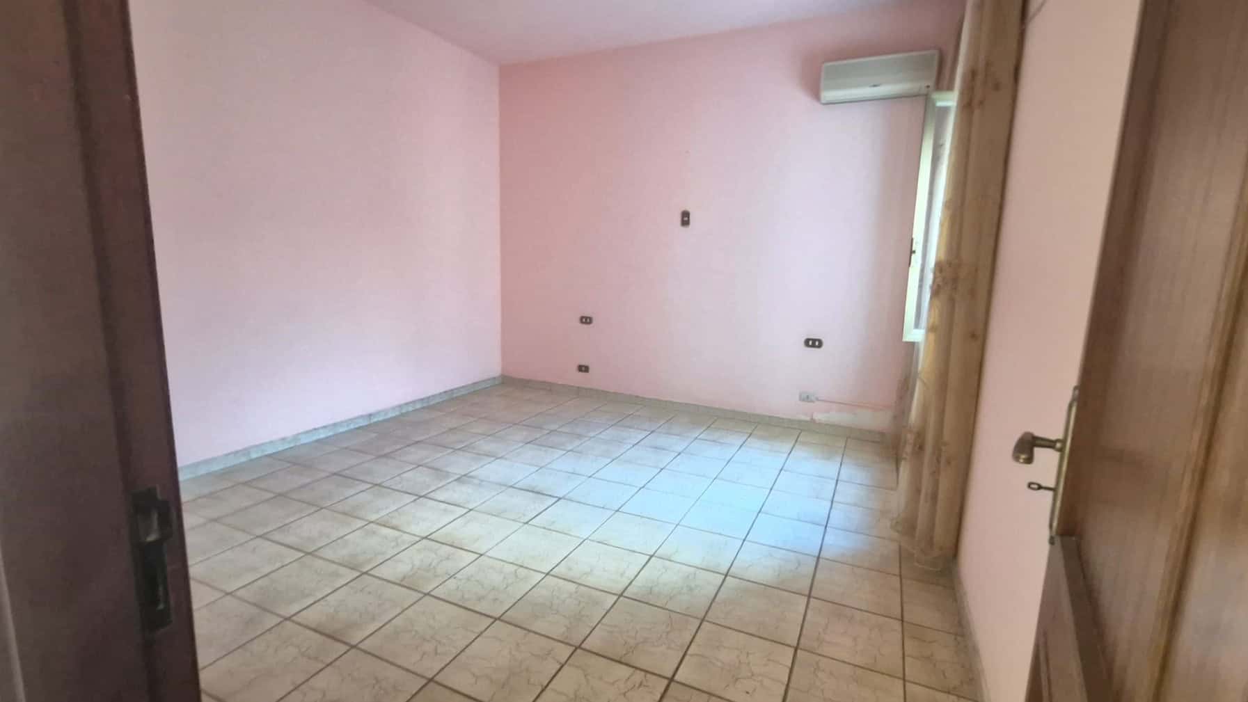 Ampia stanza vuota con pareti rosa chiaro e pavimento in piastrelle beige, illuminata da luce naturale proveniente da una finestra. Ideale come soggiorno o camera da letto, pronta per essere personali.