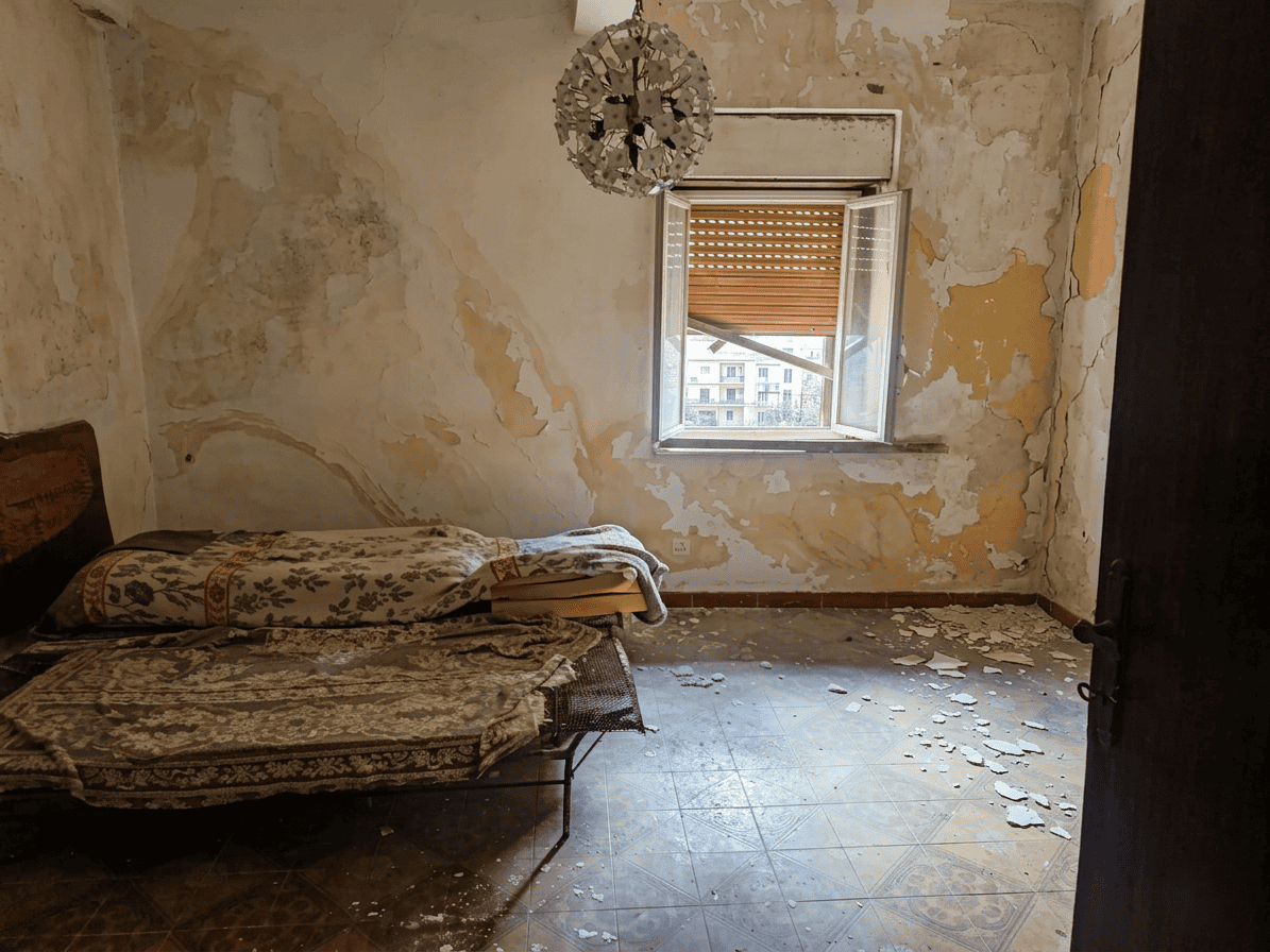 Stanza da letto abbandonata con pareti scrostate e pavimento danneggiato, luce naturale proveniente da finestra aperta, arredamento vecchio e disordine.
