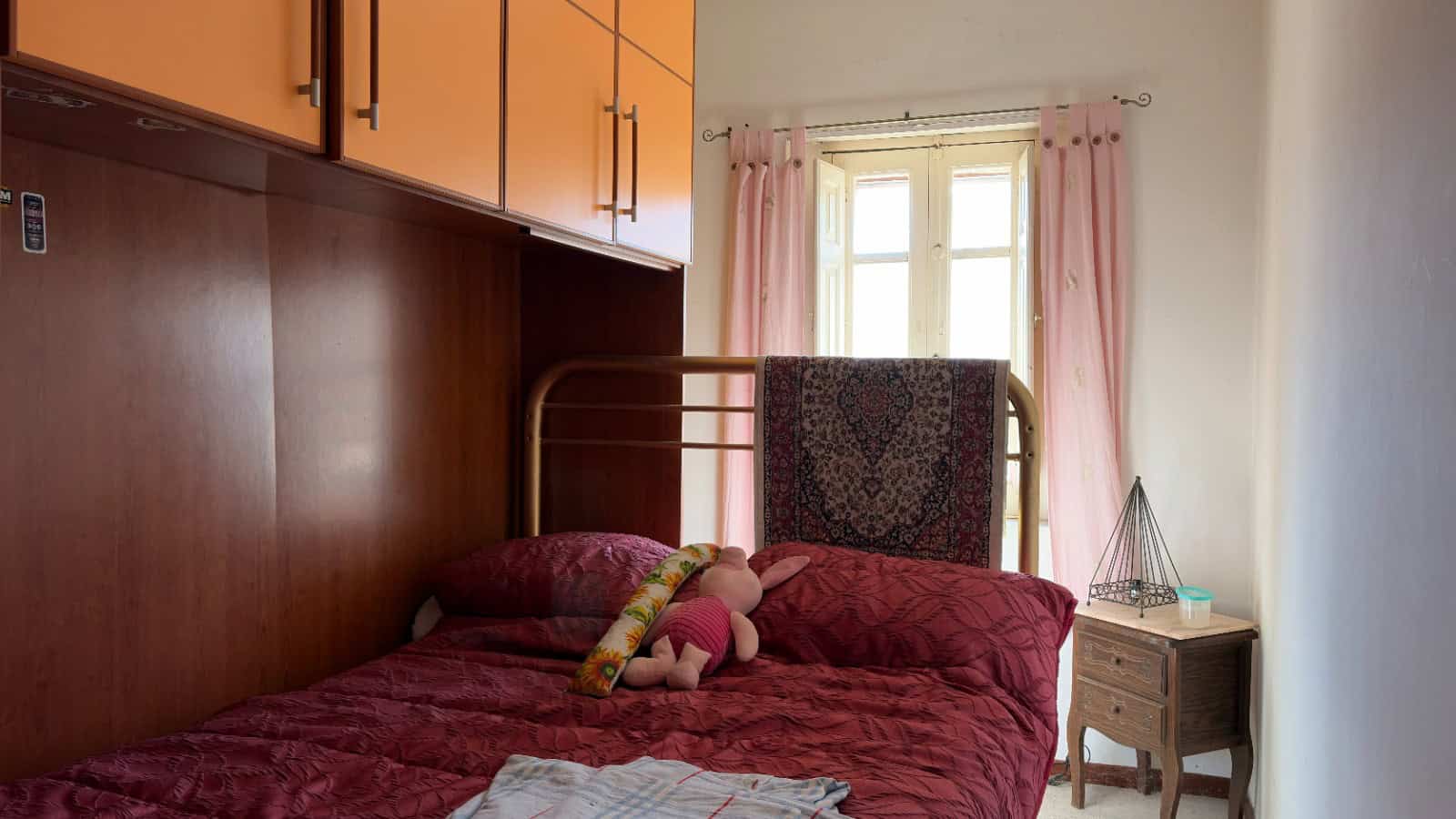 L'immagine mostra una camera da letto con letto singolo, cuscini e un peluche, illuminata da luce naturale proveniente da una finestra con tende rosa.