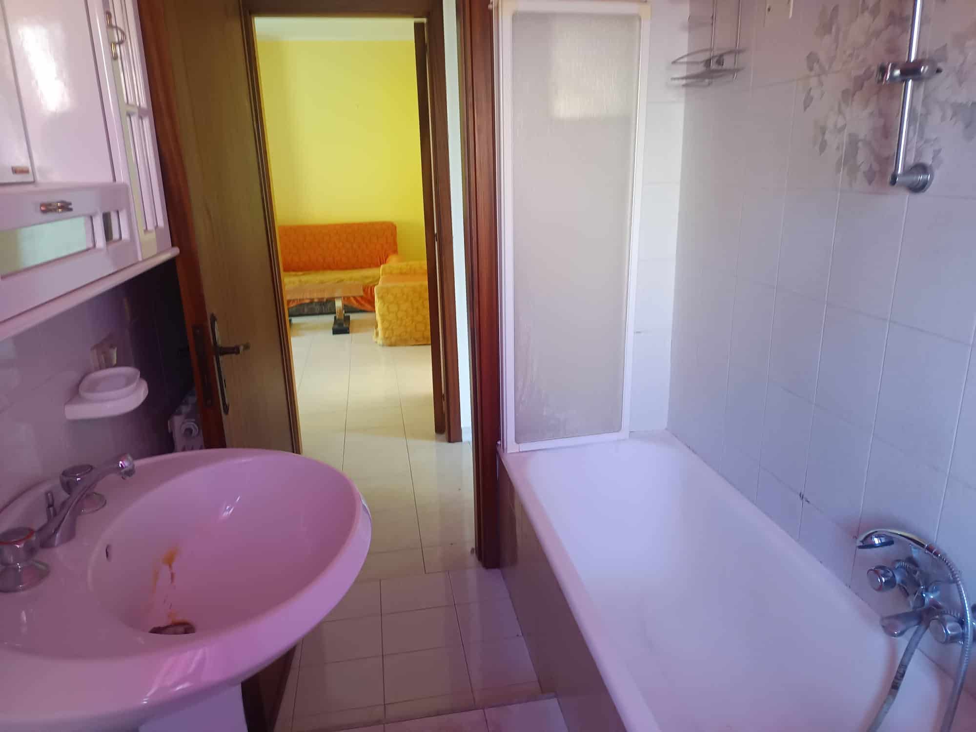 Lavabo e vasca da bagno in un bagno di appartamento con pareti bianche e piastrelle chiare, vista sulla porta che conduce a una stanza con pareti gialle e arredi colorati.