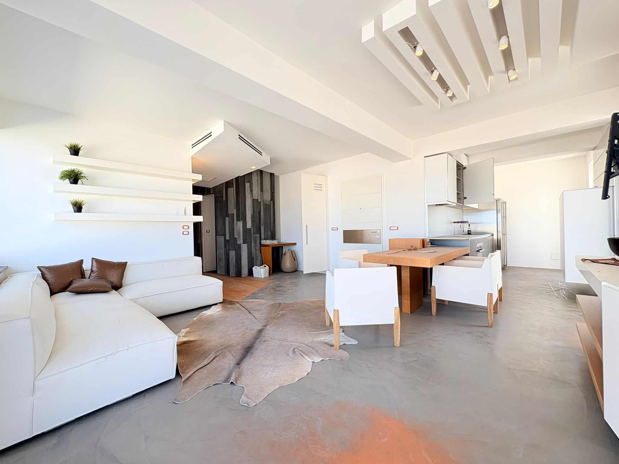 Ampia zona living moderna con divano bianco, pareti bianche e dettagli in legno, cucina aperta con isola centrale e sedie, ambiente luminoso e accogliente, ideale per vivere e ricevere ospiti.