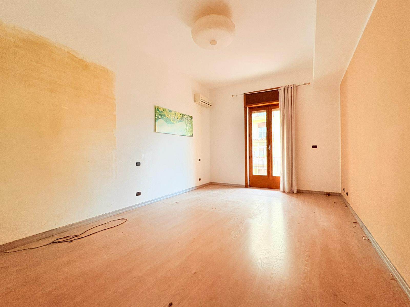 Ampio soggiorno luminoso con pareti neutre, pavimento in legno chiaro e grande finestra con accesso al balcone, ideale per vivere in comfort e stile.