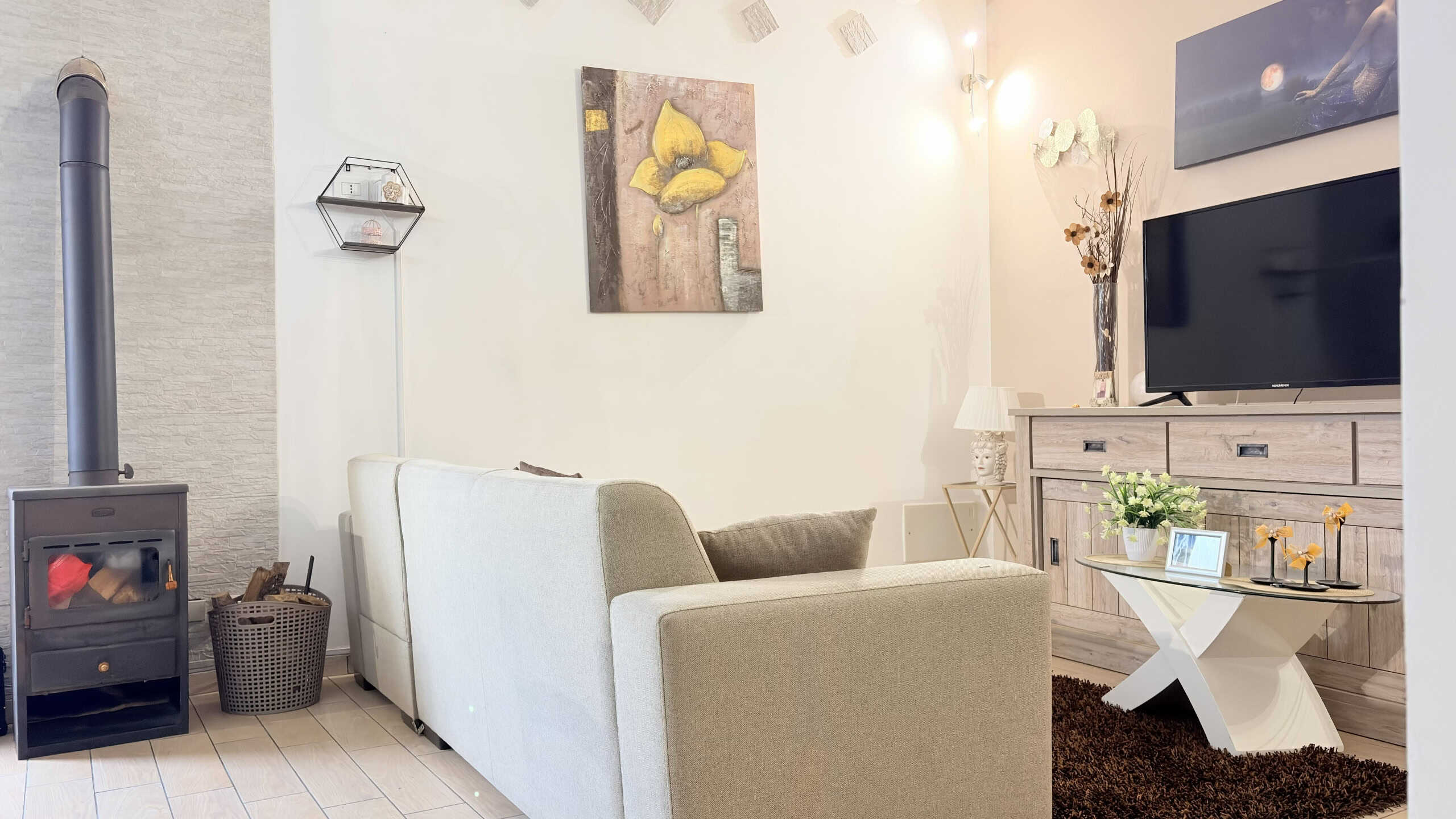 Accogliente soggiorno con divano beige, camino a legna e TV a schermo piatto, decorato con quadri e piante, ideale per momenti di relax e convivialità in casa.