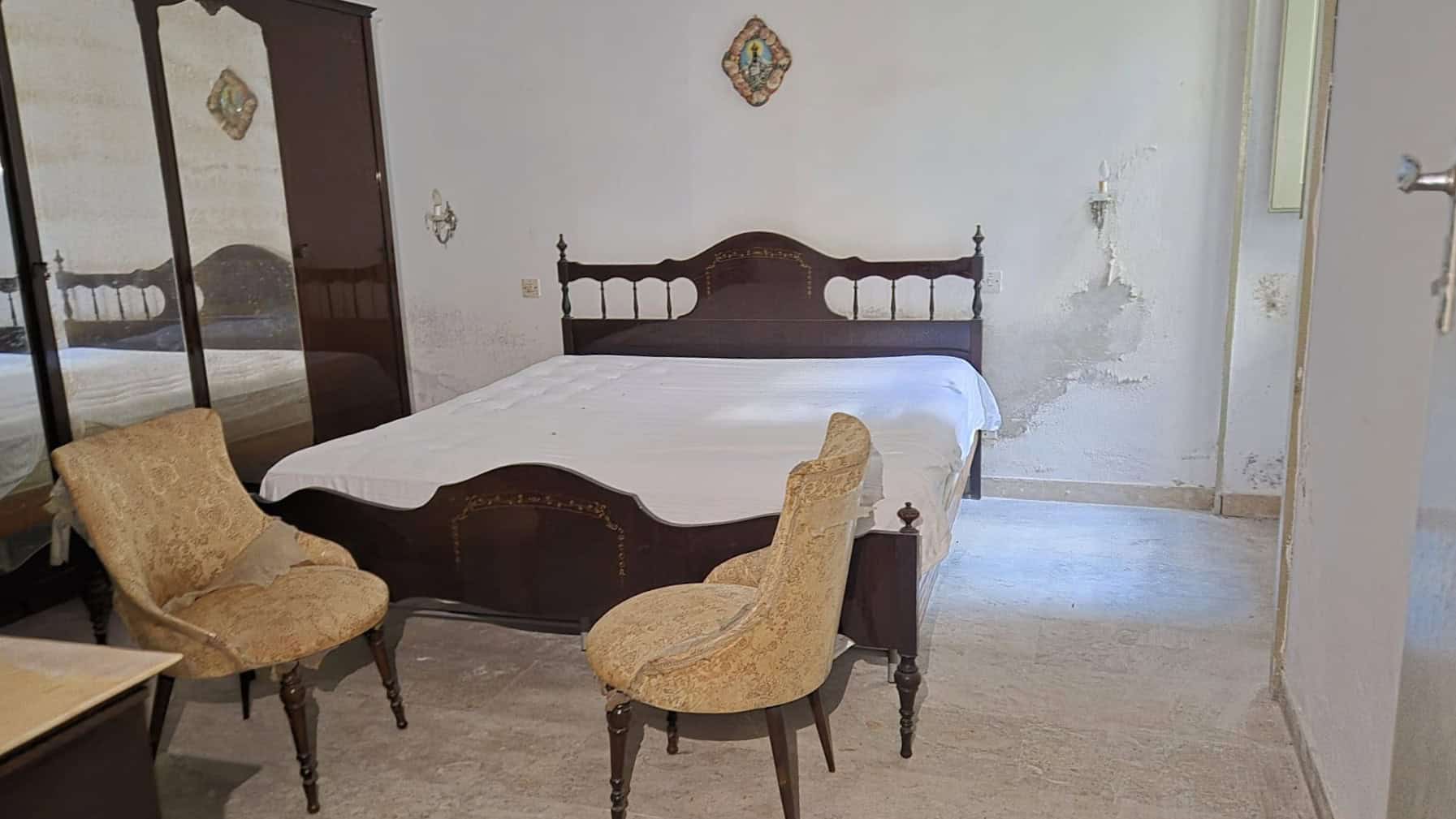 Antico letto in legno con testiera decorativa, in una stanza con pareti danneggiate e pavimento in pietra, ambiente rustico e da ristrutturare.