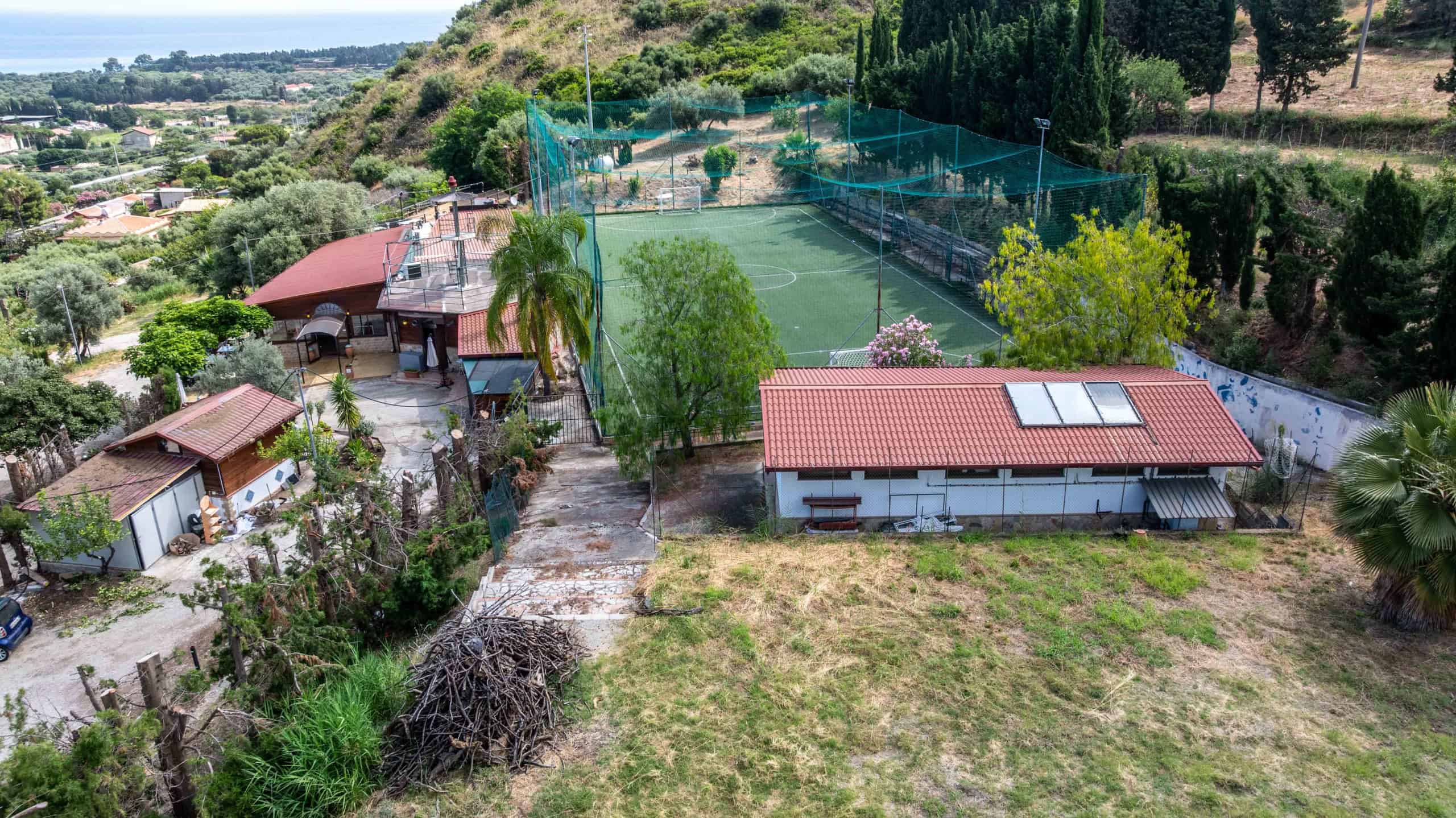 Campo da tennis all'aperto con recinzione in rete, circondato da alberi e vegetazione, in un'area residenziale tranquilla con case e colline sullo sfondo.