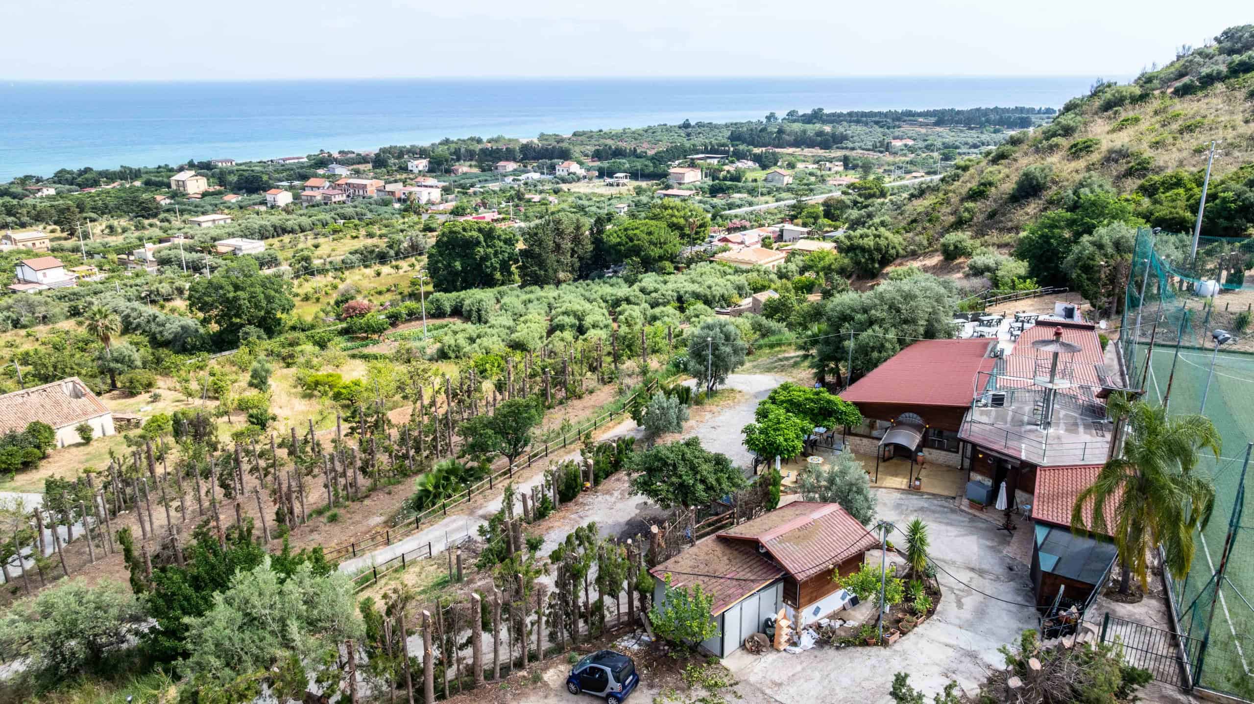 Vista panoramica di una zona residenziale con case, alberi e vegetazione, sullo sfondo il mare e l'orizzonte, con colline e cielo sereno, in un ambiente naturale e tranquillo.