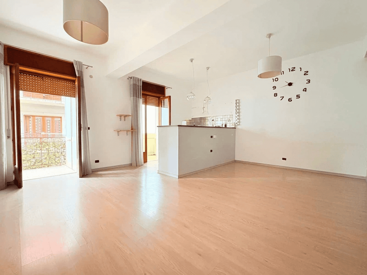 Interno luminoso e spazioso di un appartamento moderno con grandi finestre, pavimento in legno chiaro e pareti bianche, ideale per affitti o vendite immobiliari in Italia.