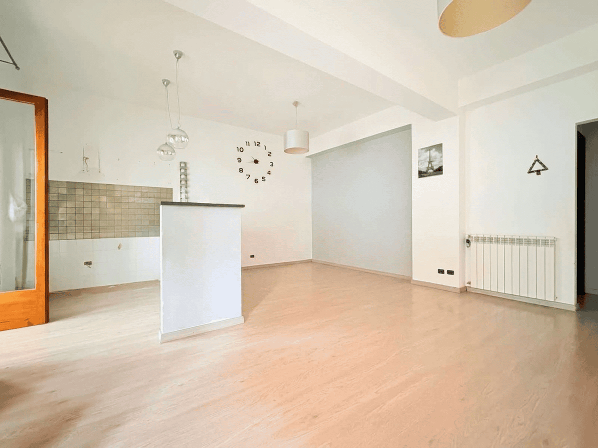 Appartamento vuoto con soggiorno luminoso, pareti bianche, pavimento in legno chiaro, cucina moderna e grande finestra, ideale per investimenti immobiliari o affitti a Milano.