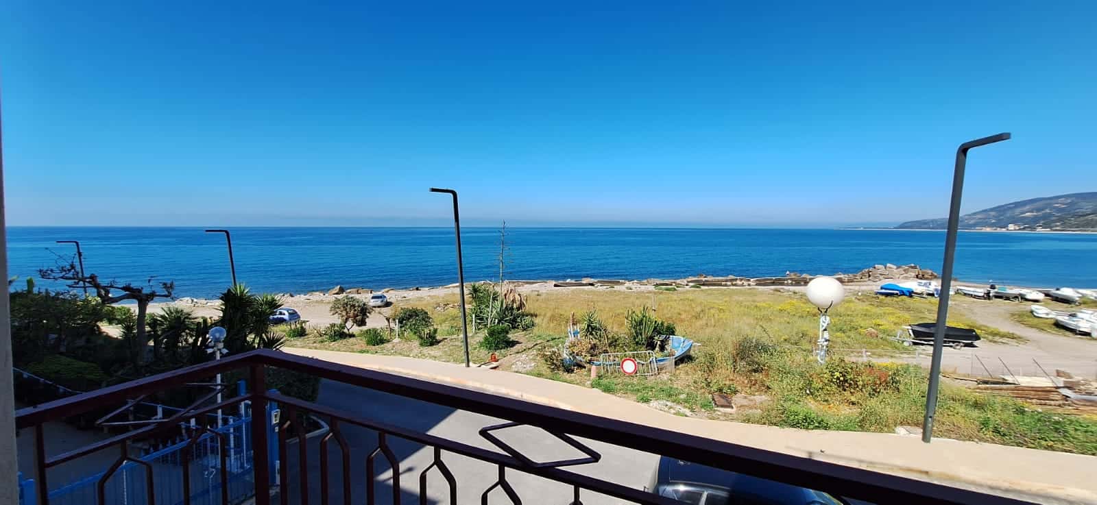 Spiaggia con vista mare, balcone con ringhiera in ferro, lampioni e vegetazione, perfetta per case vacanze o immobili fronte mare a NextCasa.