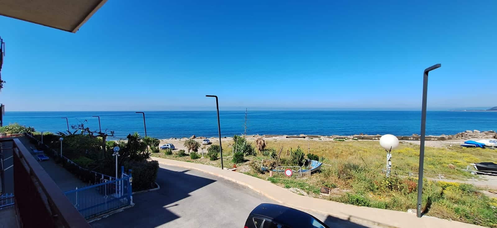 Vista panoramica sul mare con spiaggia e strada, illuminata dal sole, in un ambiente residenziale vicino alla costa, ideale per case vacanza o proprietà fronte mare.