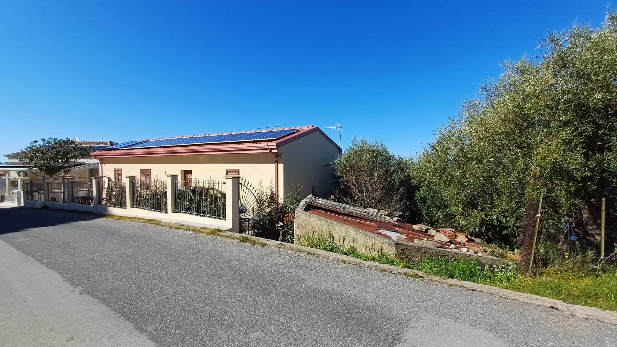 Ampia casa residenziale con giardino e strada asfaltata, sotto un cielo sereno, circondata da vegetazione e alberi, in un contesto tranquillo e ben mantenuto.