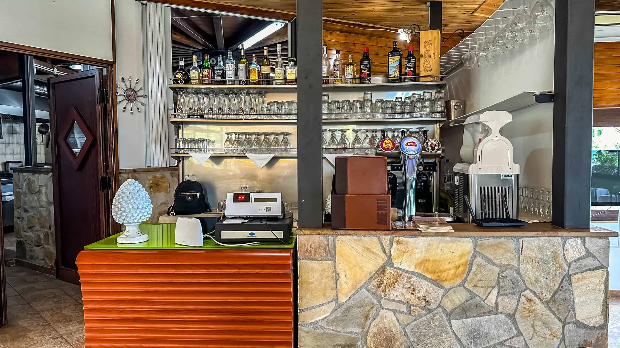 Bar in stile rustico con scaffali di vetro pieni di bottiglie e bicchieri, arredamento caldo in legno e pietra, ideale per ambienti di ristorazione o bar accoglienti.