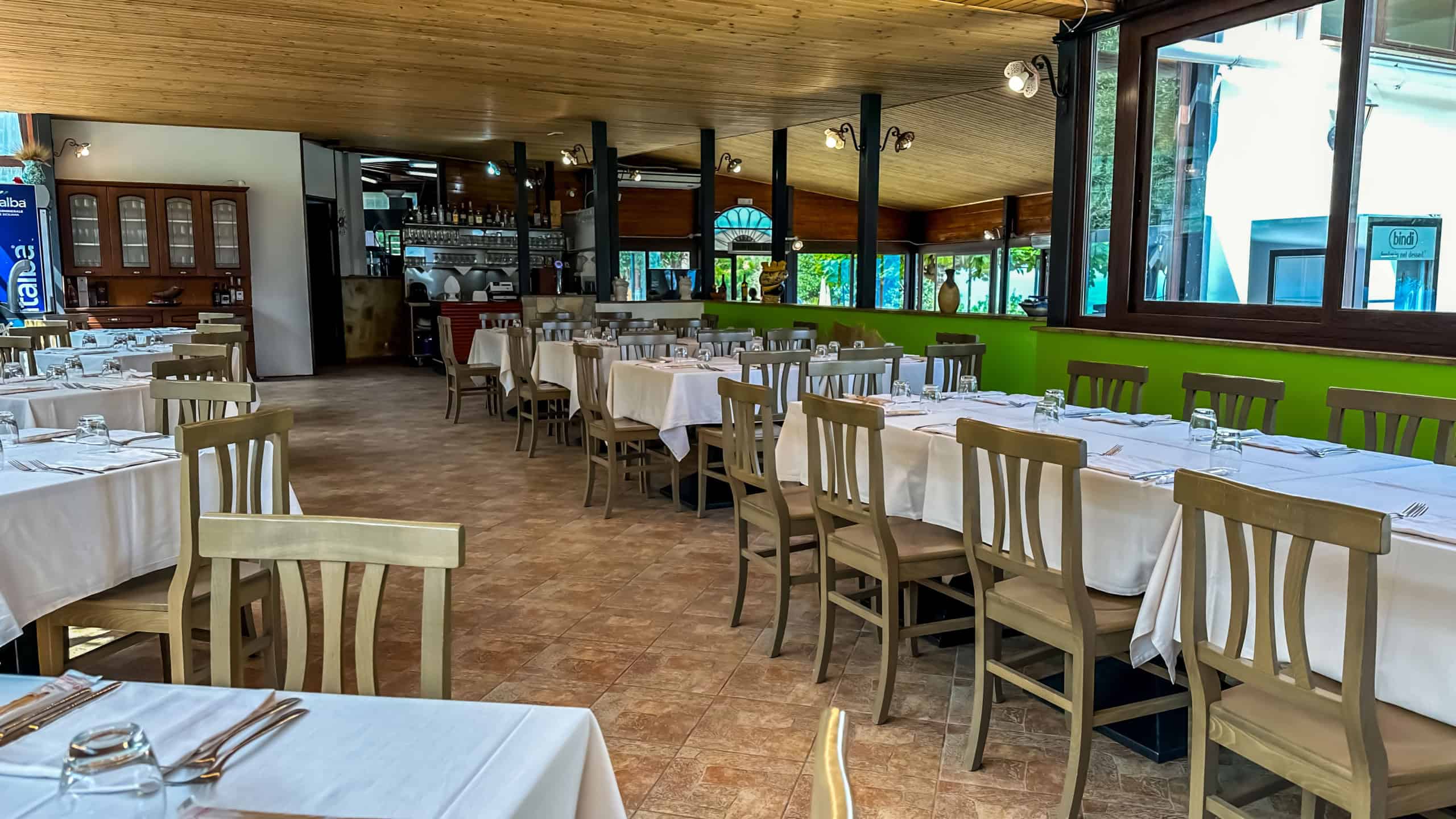 Tavolo elegante in ristorante con sedie in legno, tovaglie bianche e ambiente accogliente con grandi finestre e illuminazione calda, ideale per cene e eventi speciali.