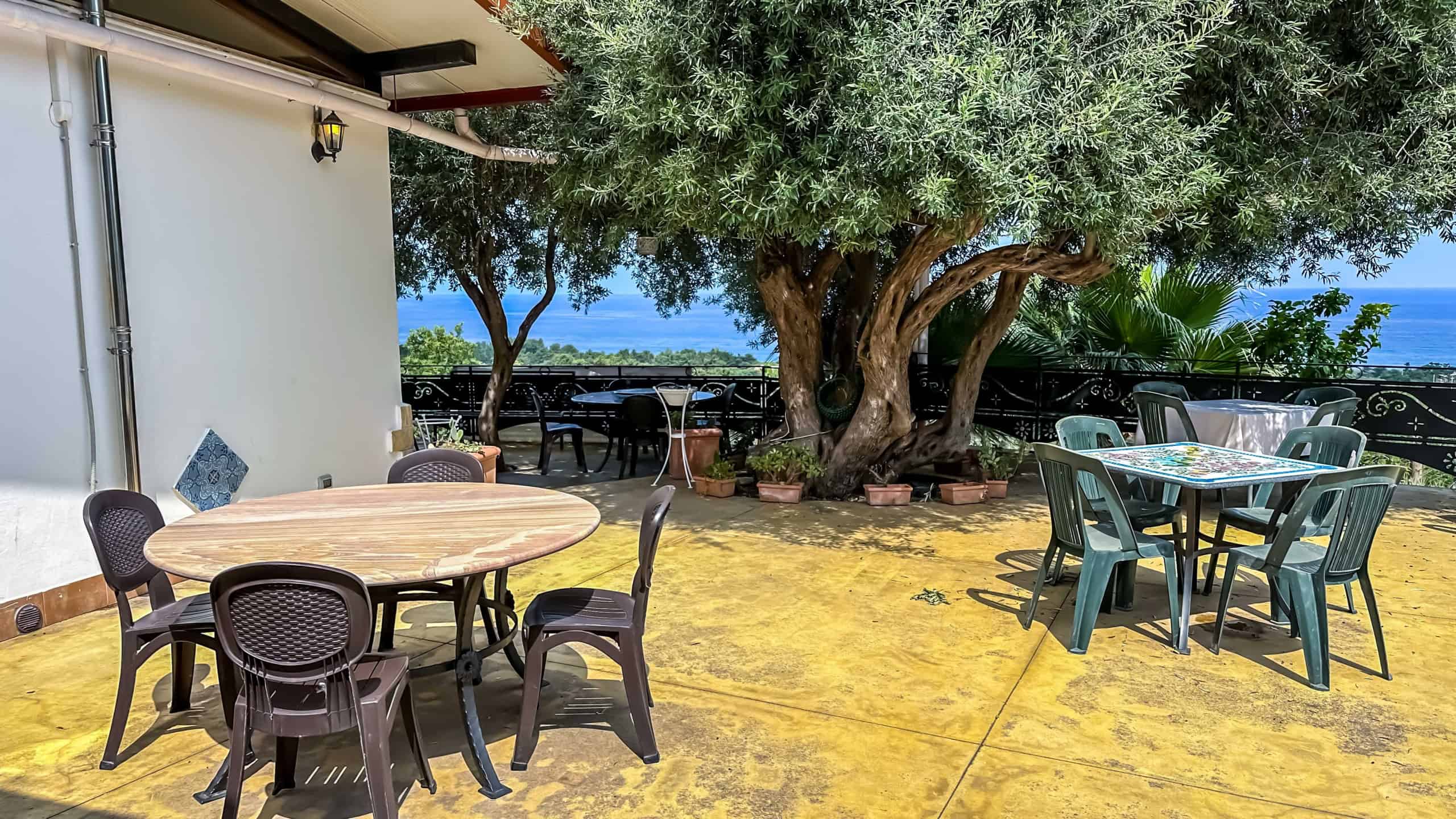 Ampia terrazza con tavoli e sedie, vista mare e grande albero ombreggiante, ideale per relax e cene all'aperto in un ambiente tranquillo e suggestivo.